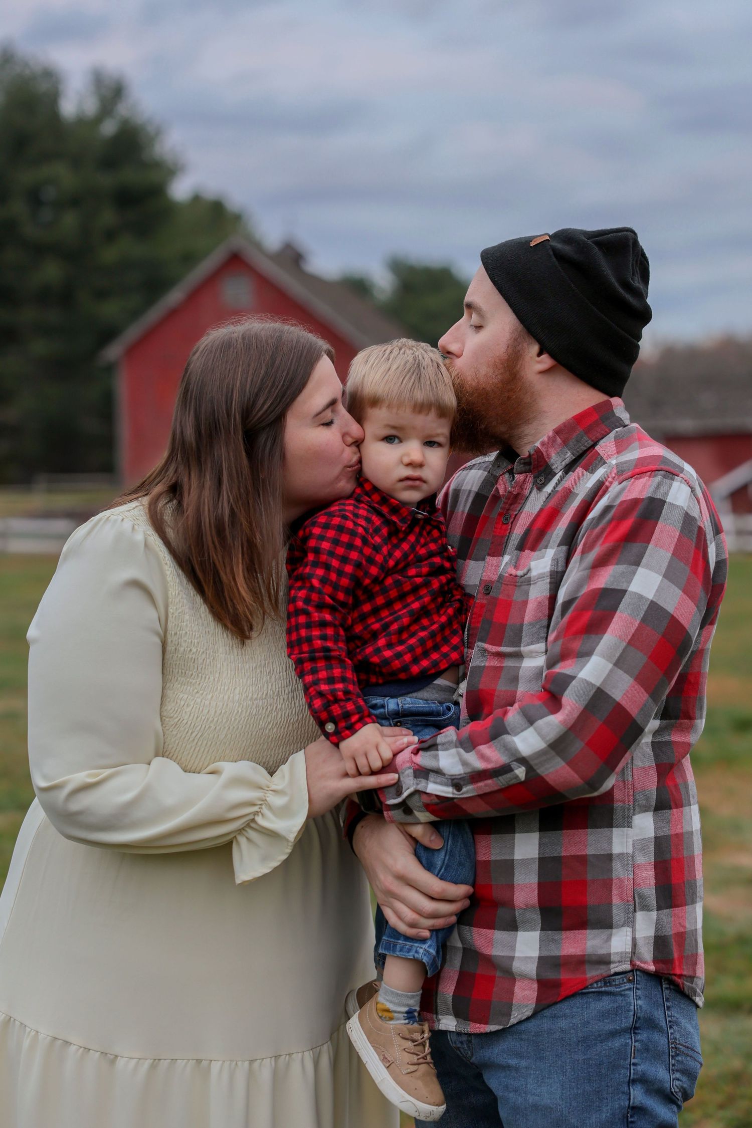 Parents in coordinating outfits kissing toddler son on cheeks simultaneously with red barn in background.