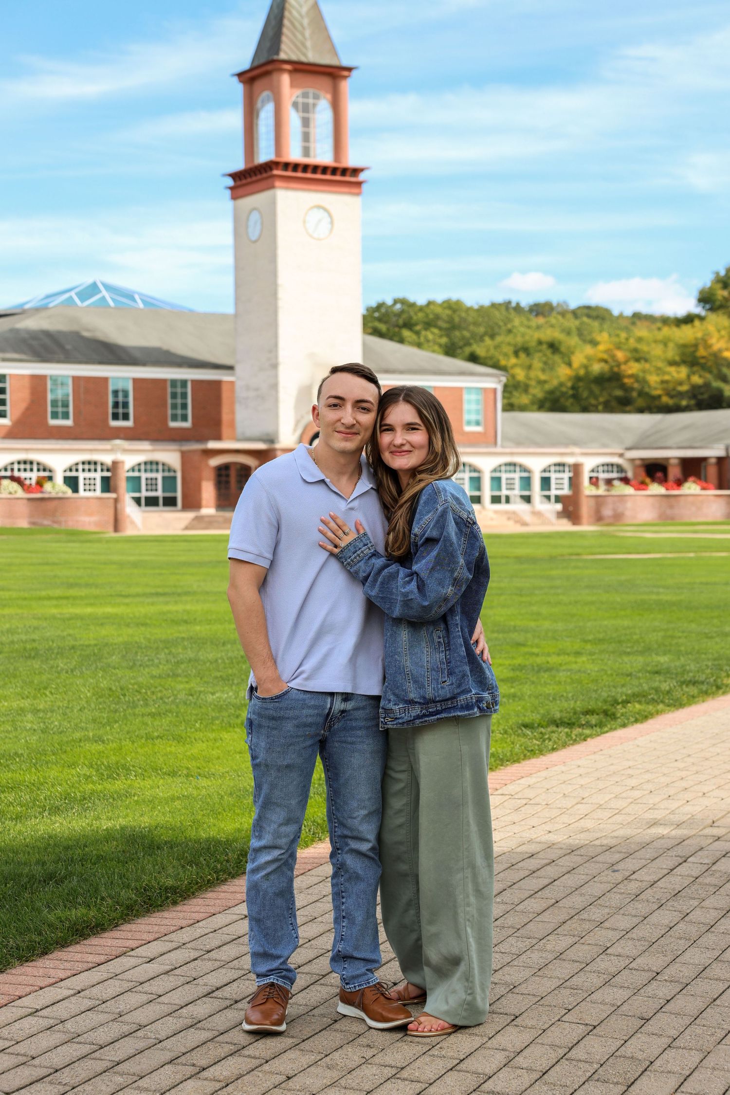 Young couple embracing and smiling in front of a brick clock tower on a college campus on a sunny day.
