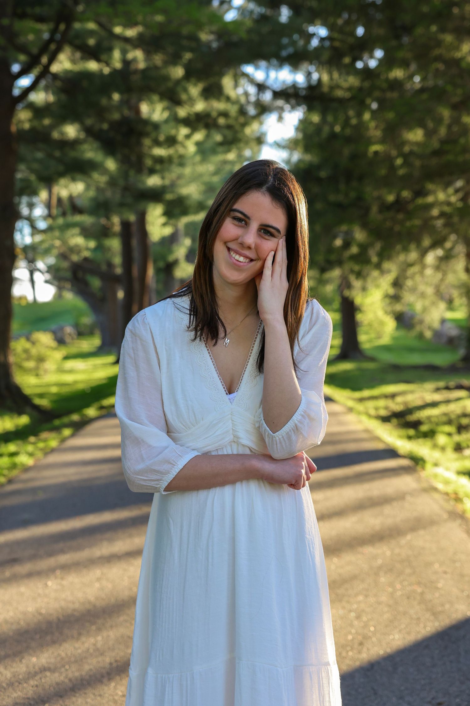 Smiling brunette woman in white dress standing on tree-lined road in golden light.