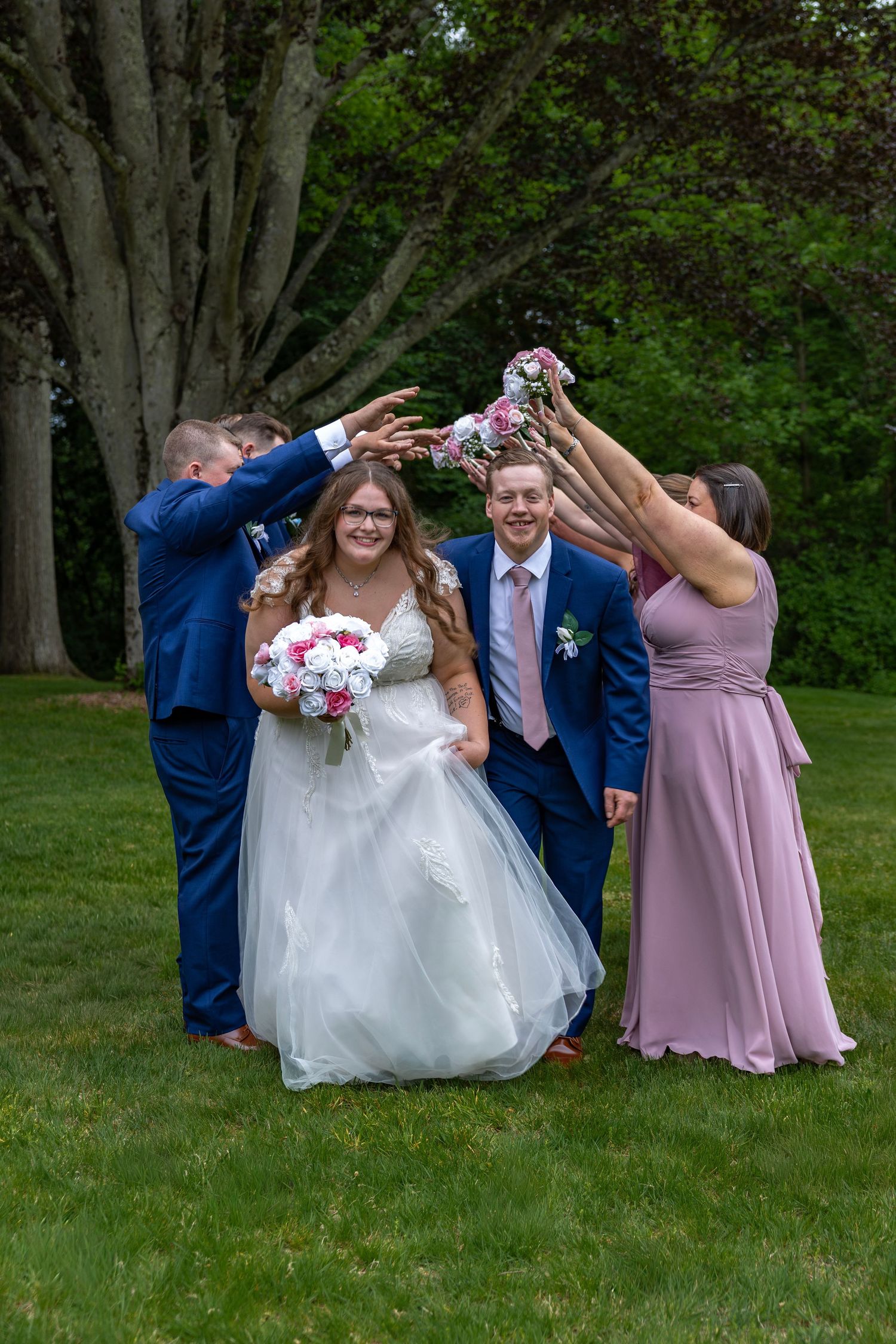 Wedding party creates arch with bouquets as bride and groom walk through on green lawn outdoors.