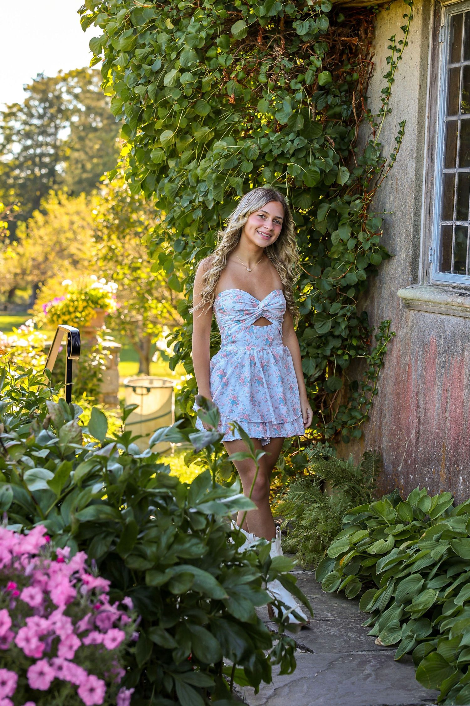 Blonde young woman in floral sundress standing on garden path beside ivy-covered stone building with pink flowers.