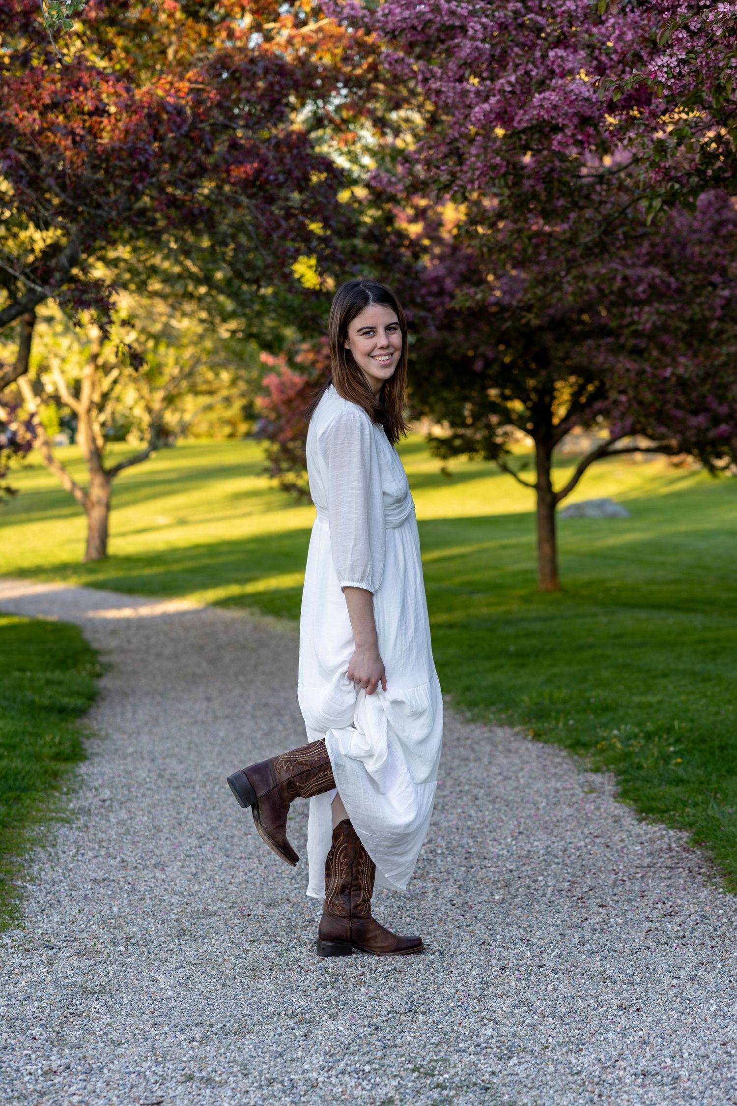 Woman in white dress and cowboy boots posing on path near pink flowering trees.
