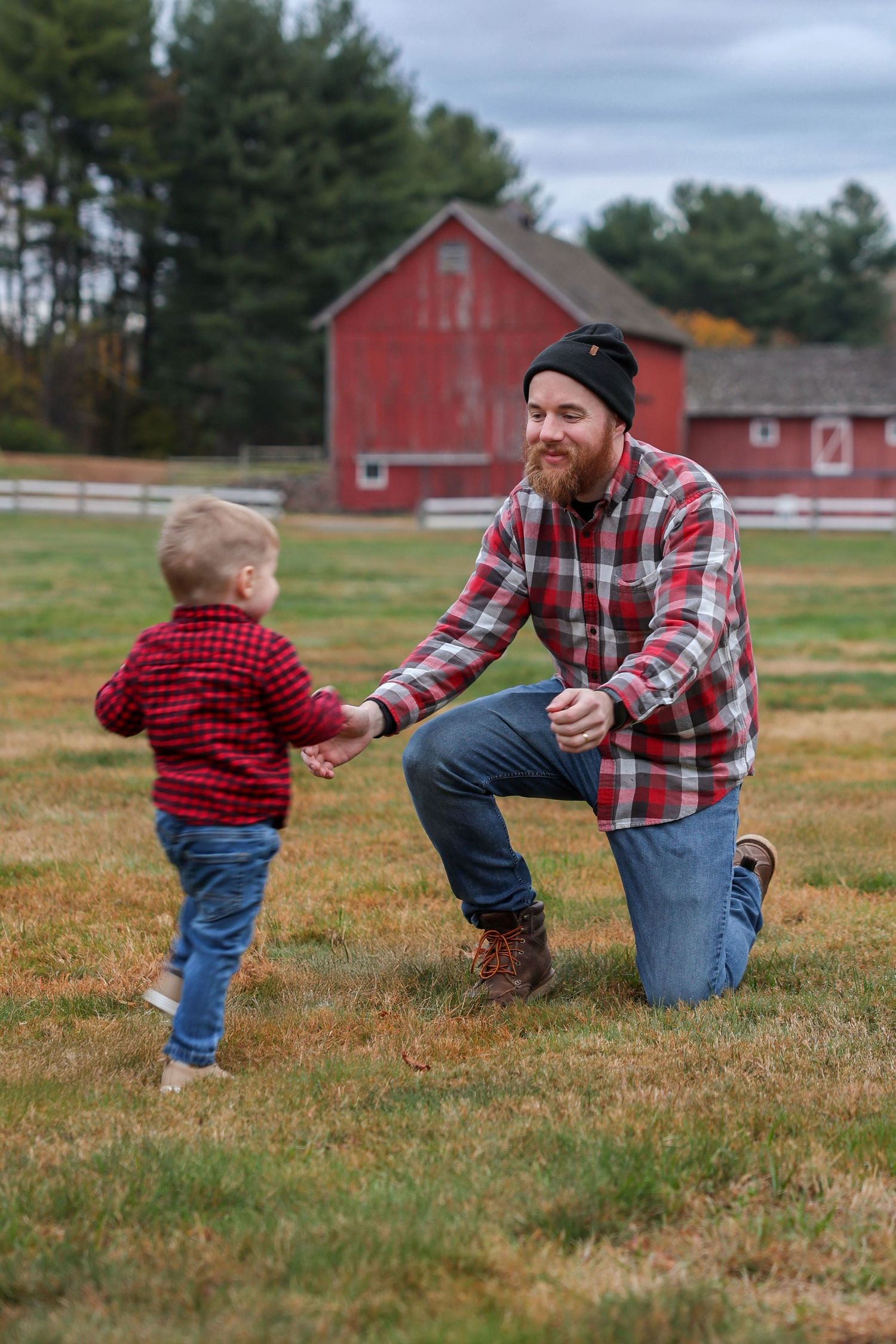 Bearded father in red plaid shirt kneeling to hold hands with toddler son on farm field.
