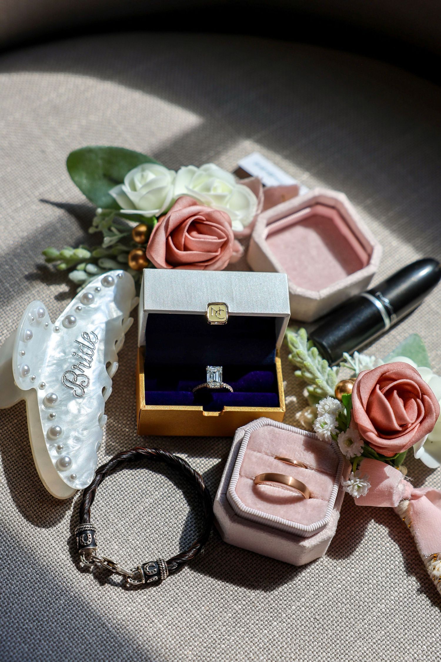 Wedding flat lay showcasing engagement ring, gold wedding band, bracelet, lipstick, and floral boutonnieres.