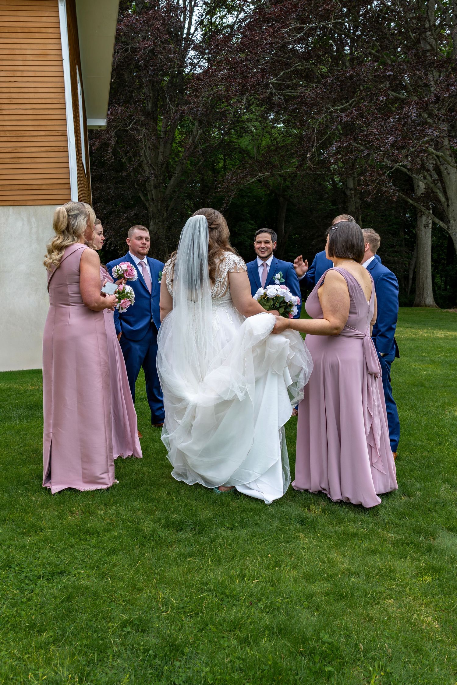 Wedding party gathers on green lawn, bride in white gown with veil surrounded by bridesmaids in pink and groomsmen in navy.