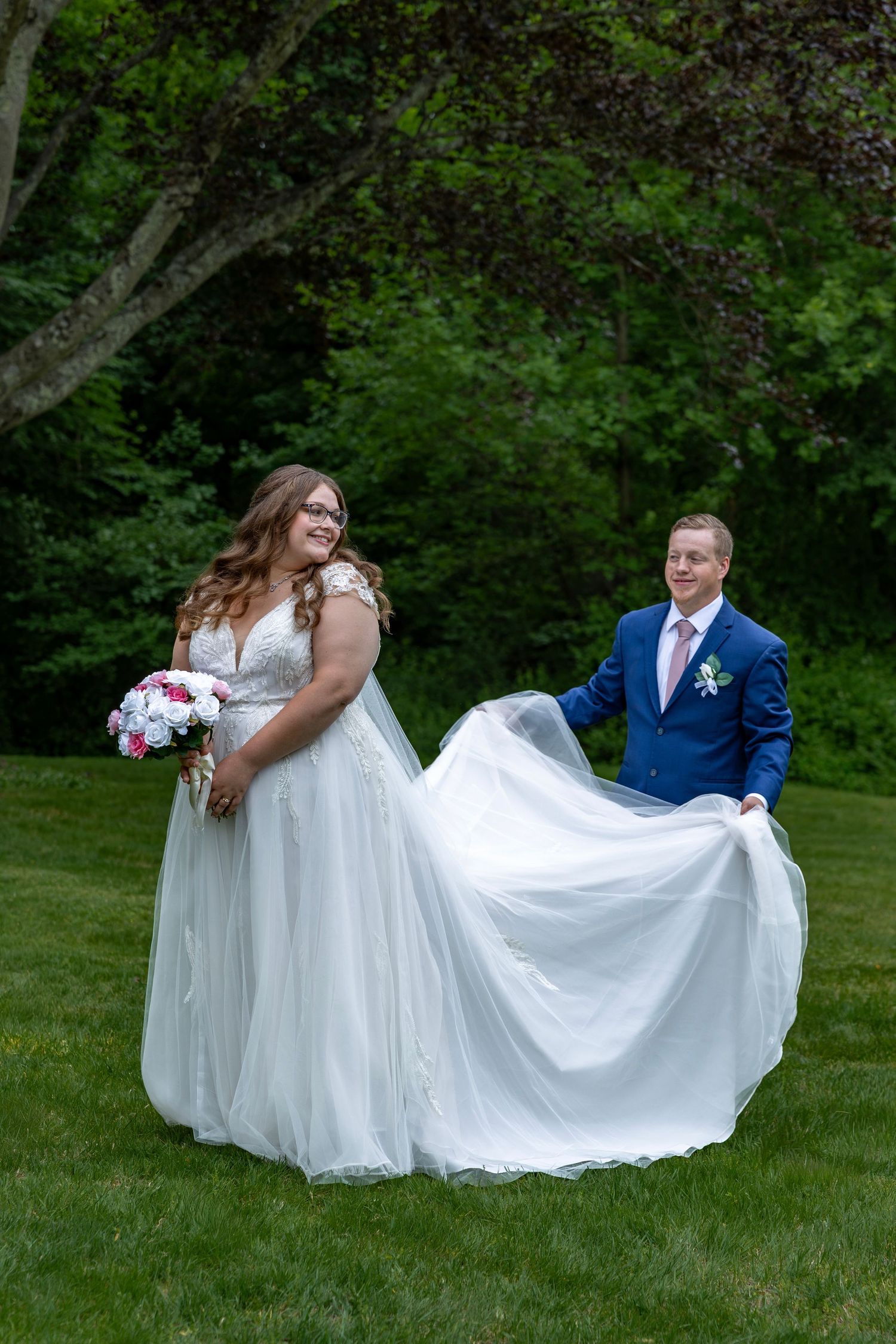 Groom in blue suit playfully fans out bride's white tulle wedding gown train on green grass outdoor.