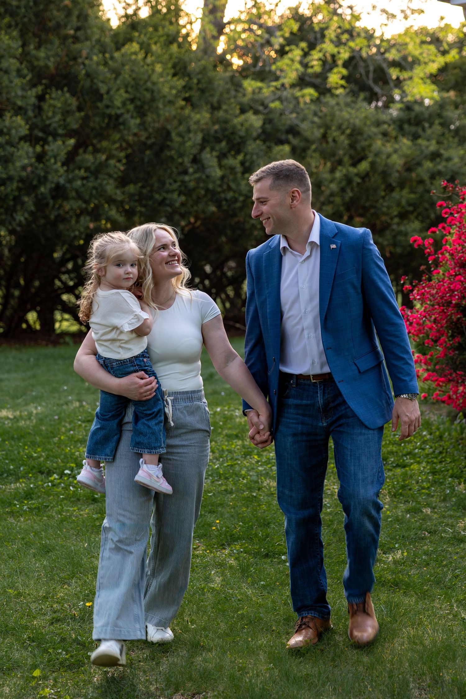 Happy family walking hand in hand through lush green garden, mother carrying toddler daughter at sunset.