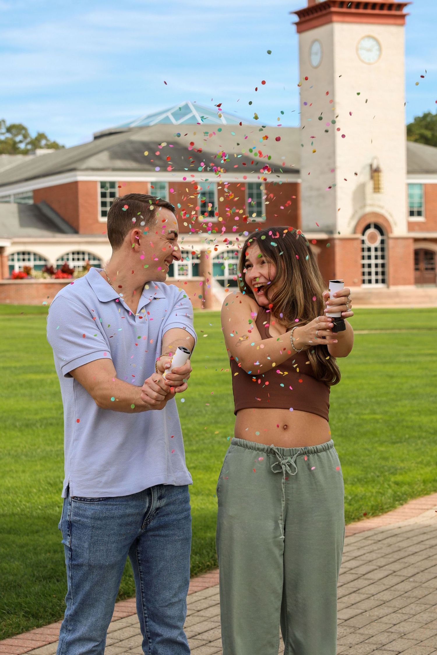 Excited couple popping confetti cannons outdoors on a campus lawn with a brick clock tower behind them.