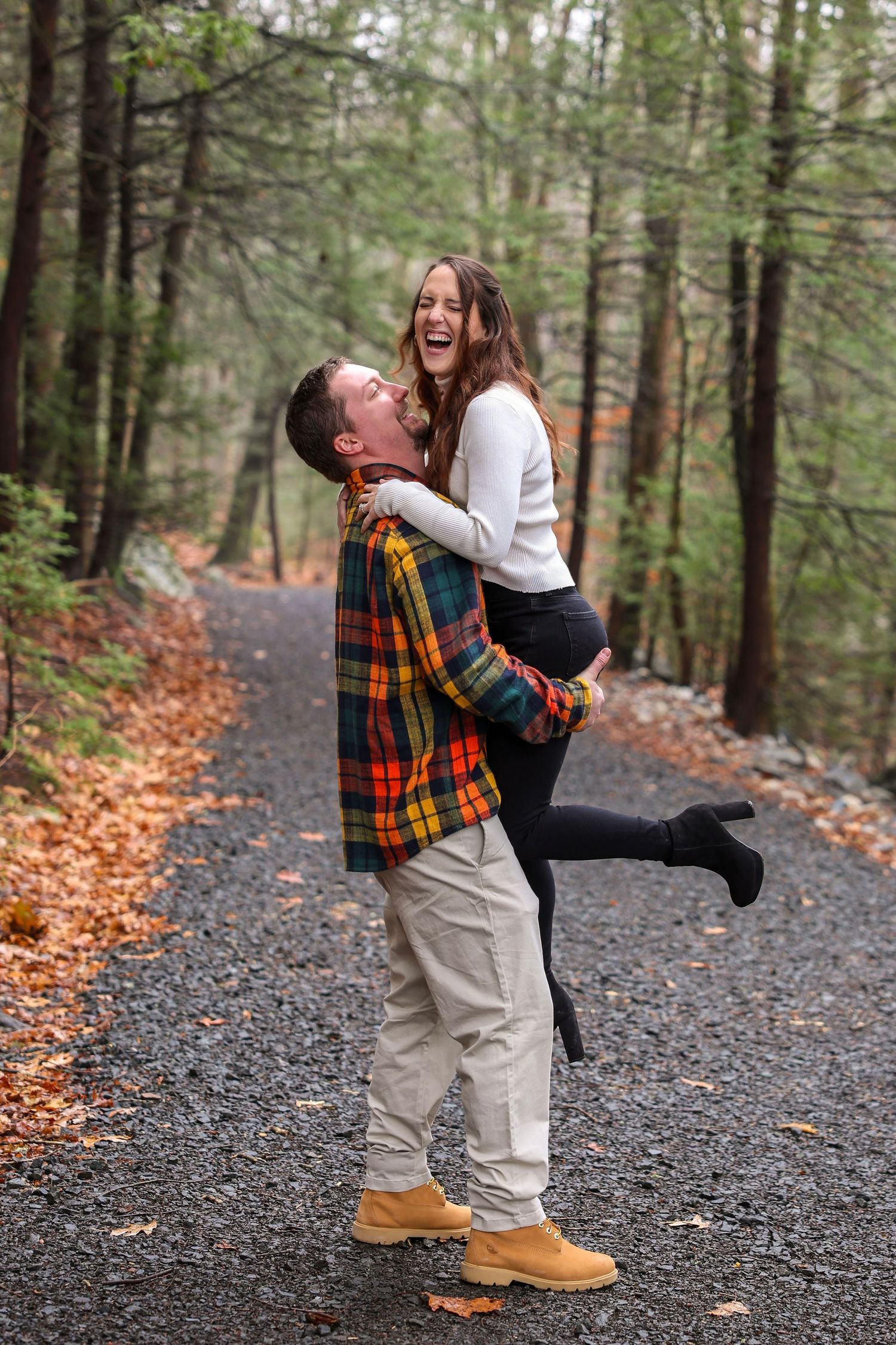A man lifts a laughing woman on a misty gravel forest road surrounded by colorful autumn foliage.