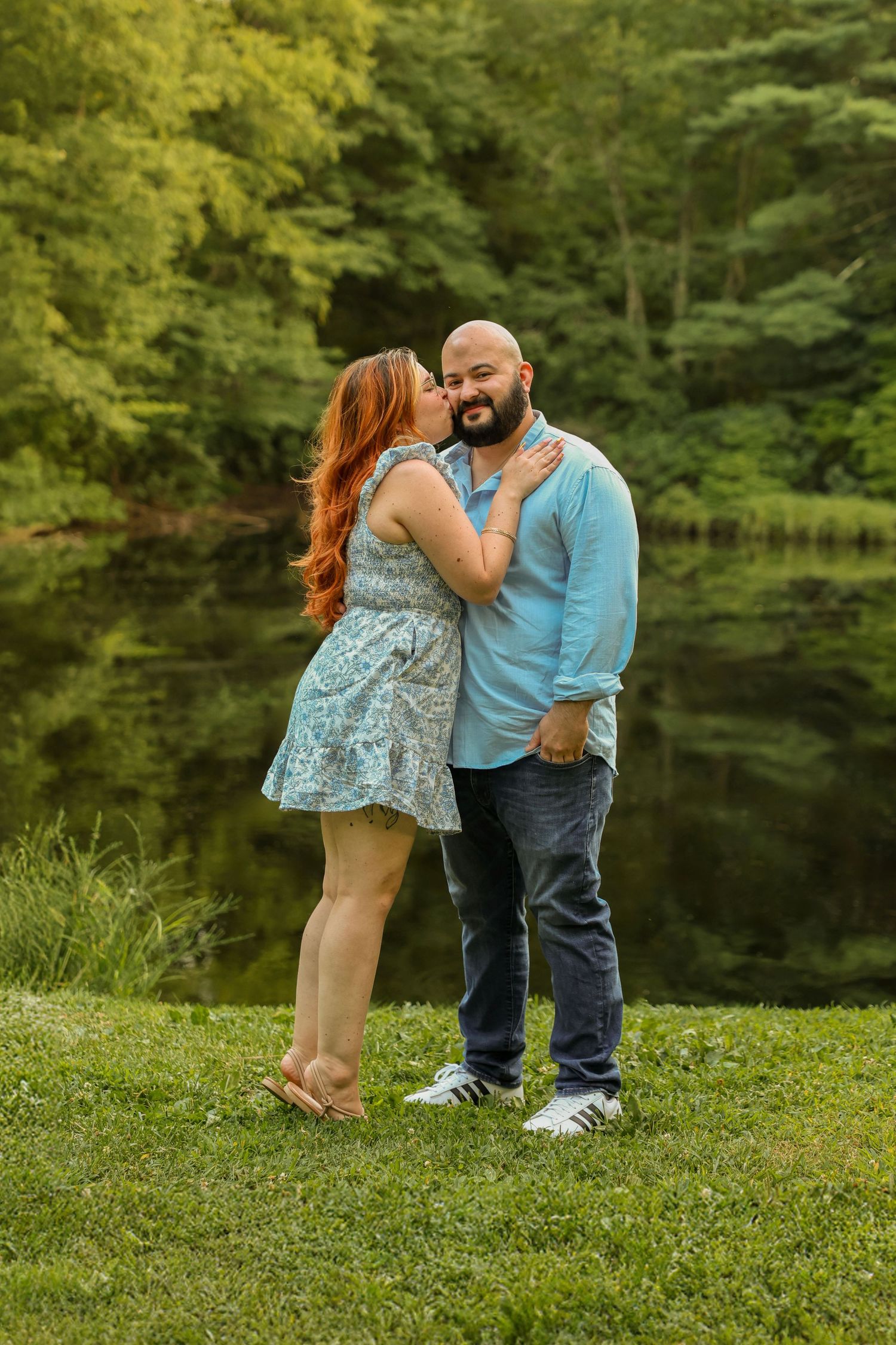 Couple sharing a kiss by a serene pond surrounded by lush green trees during a romantic outdoor photo session.