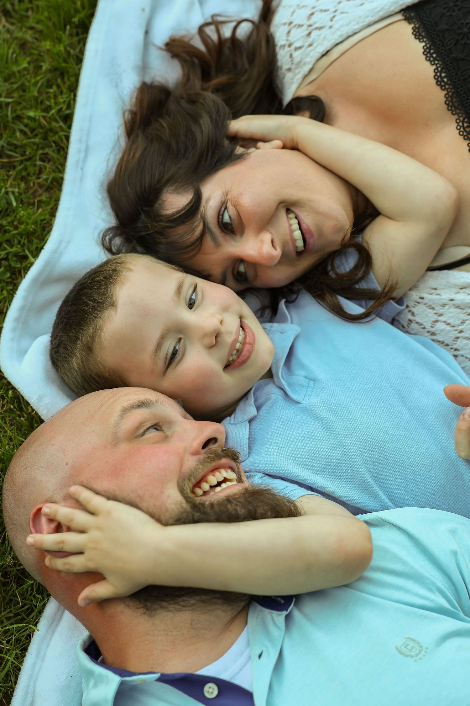 A cheerful family of three lies on a blanket in the grass, laughing and enjoying time together.