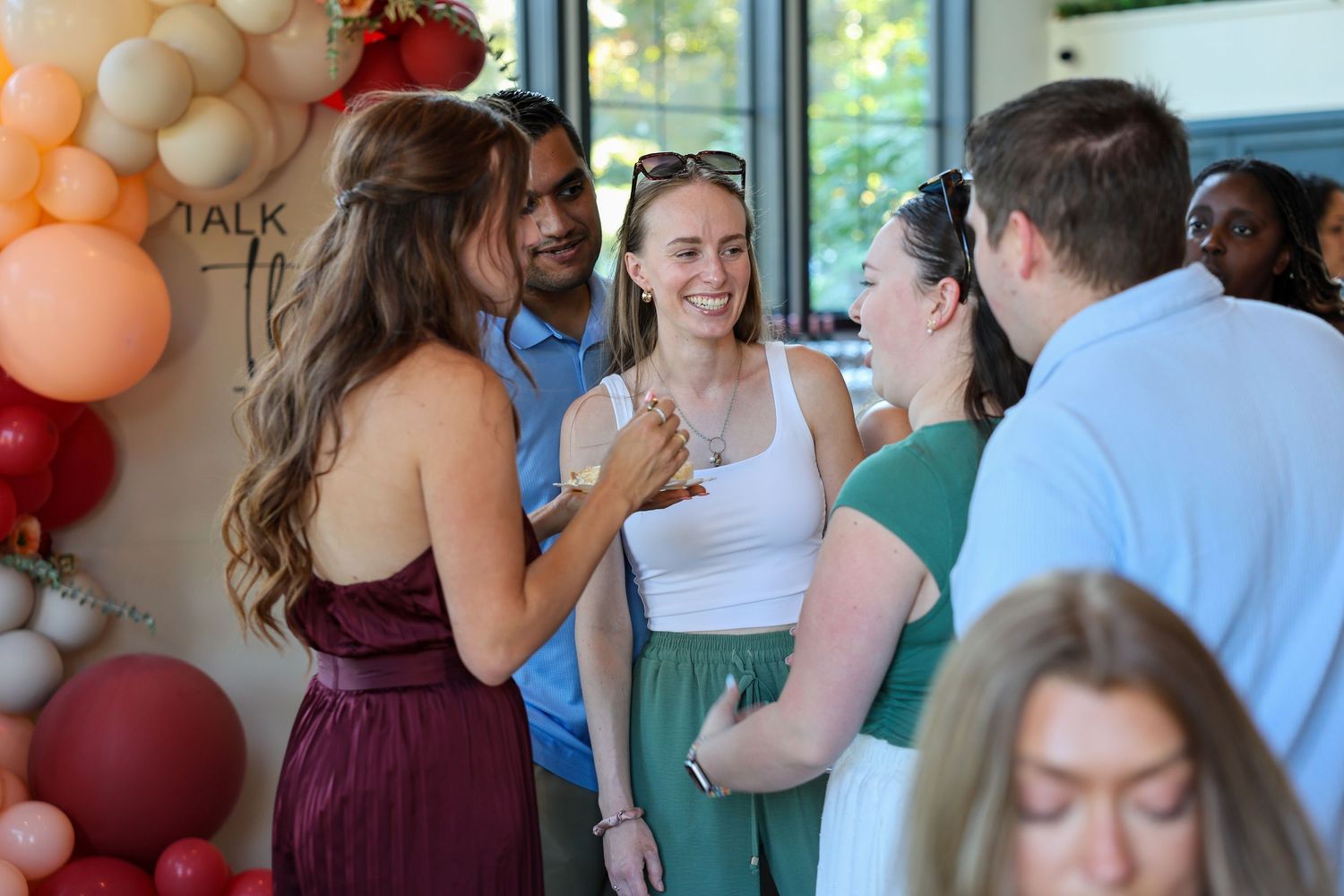 Group of guests mingling and laughing near balloon arch at an indoor celebration event.
