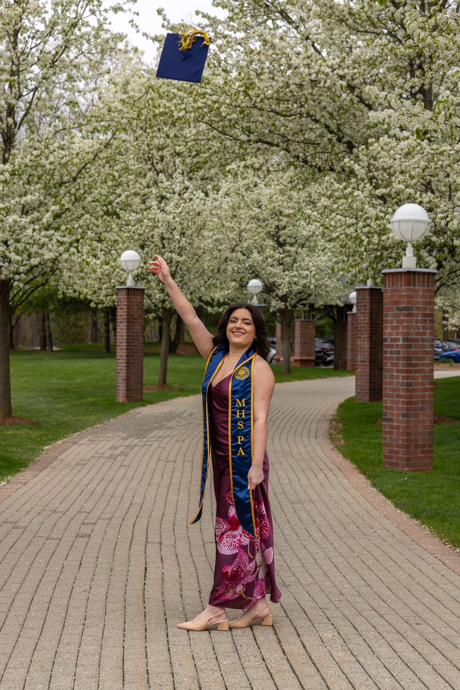 Joyful female graduate tossing cap in air on brick pathway lined with white flowering trees.