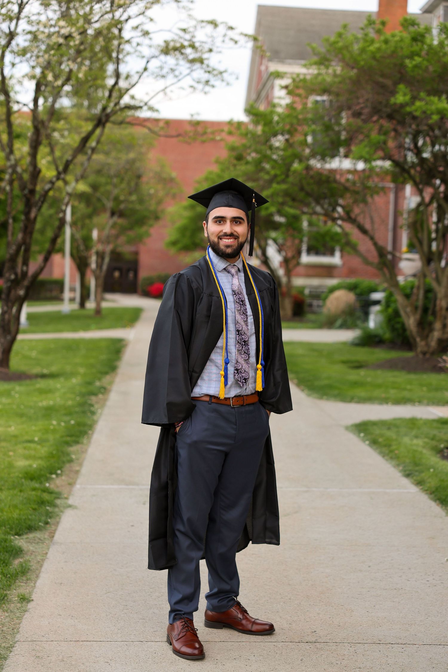 Smiling male graduate in black cap and gown with gold stole standing on campus pathway.