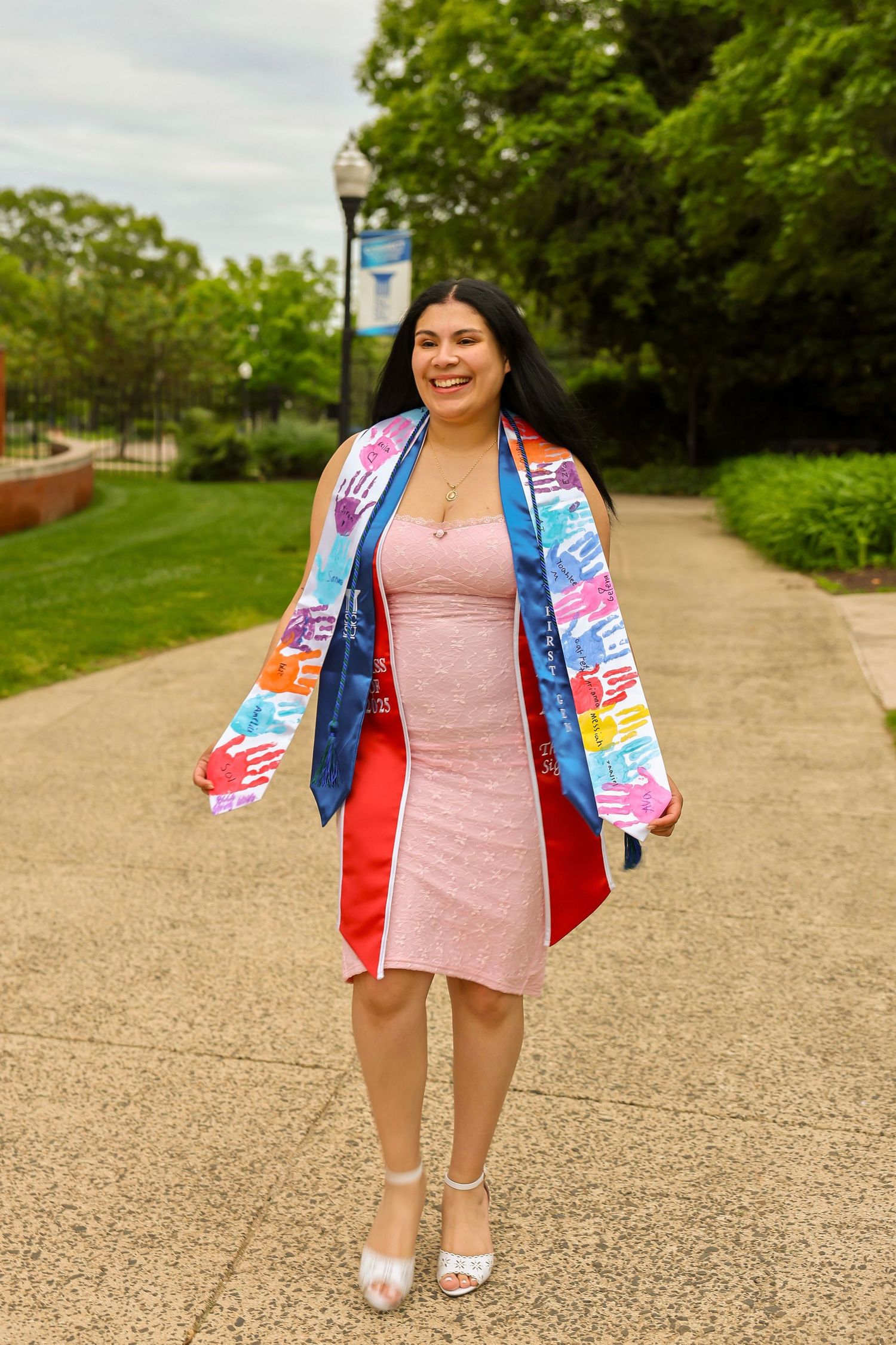 Happy graduate in pink dress wearing colorful graduation stole walking on campus pathway among green trees.
