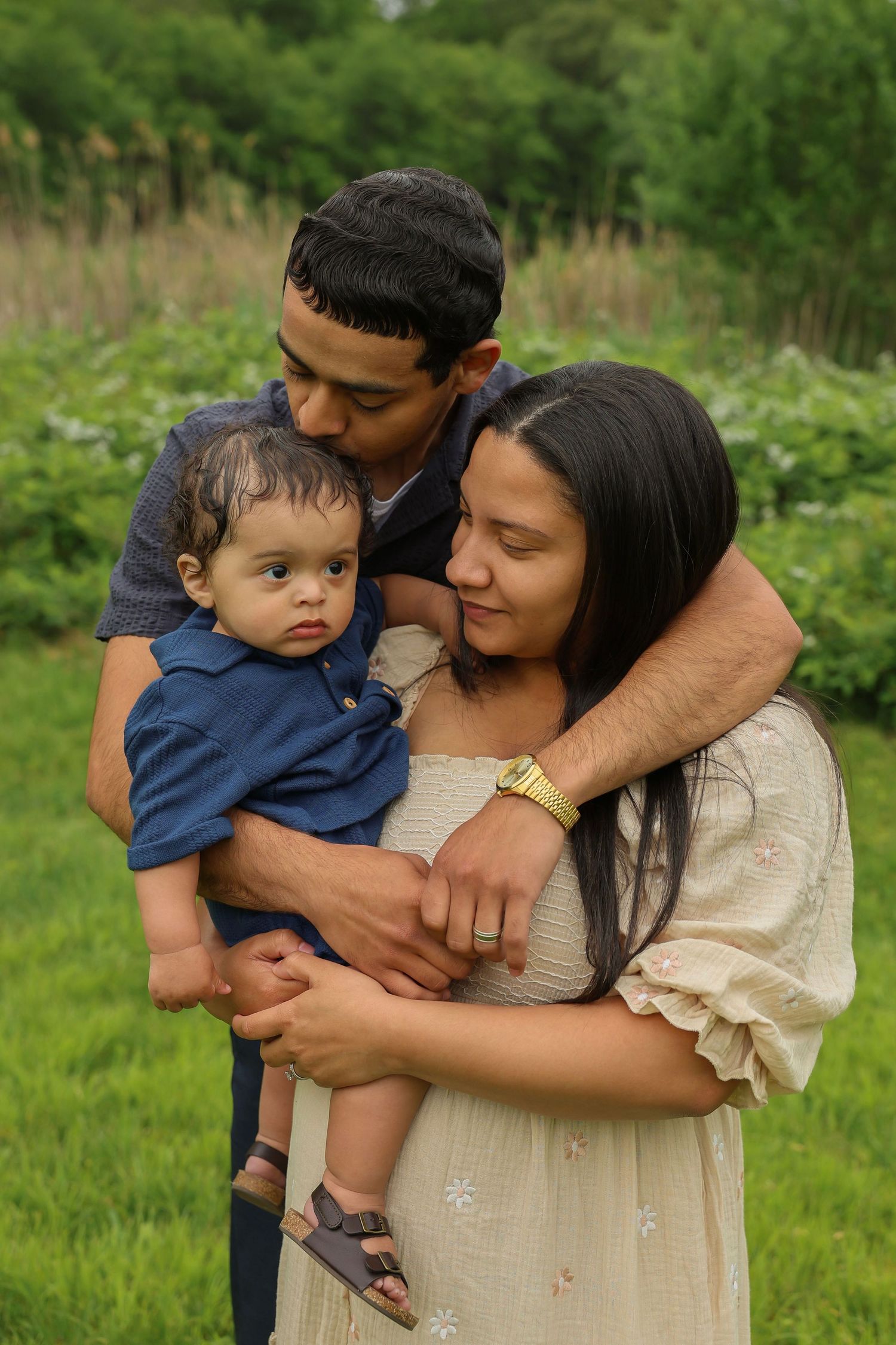 Father kisses baby's head while mother holds child in lush green outdoor setting.