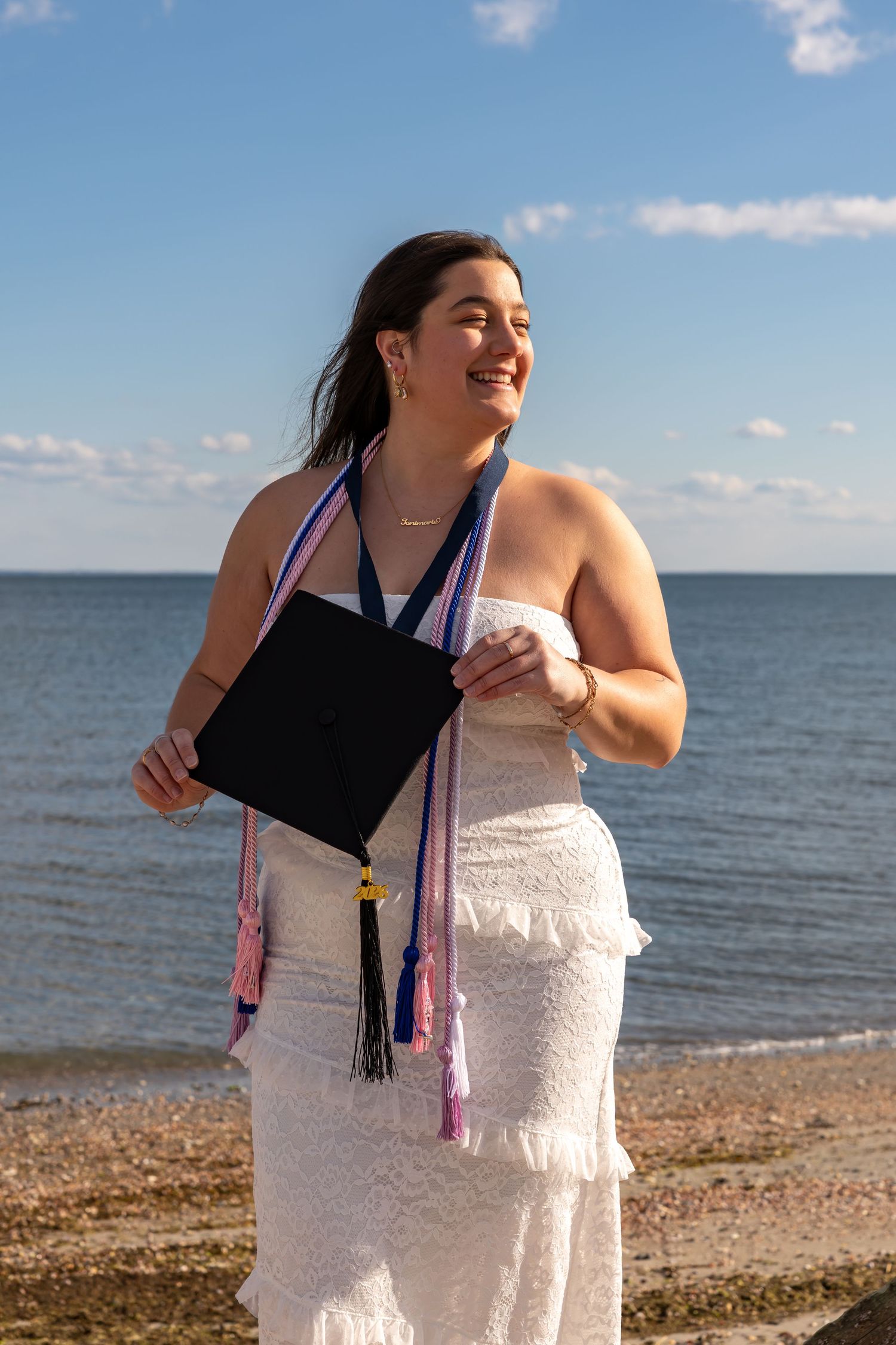 Happy young woman in white dress holding graduation cap on a sunny beach with blue sky.