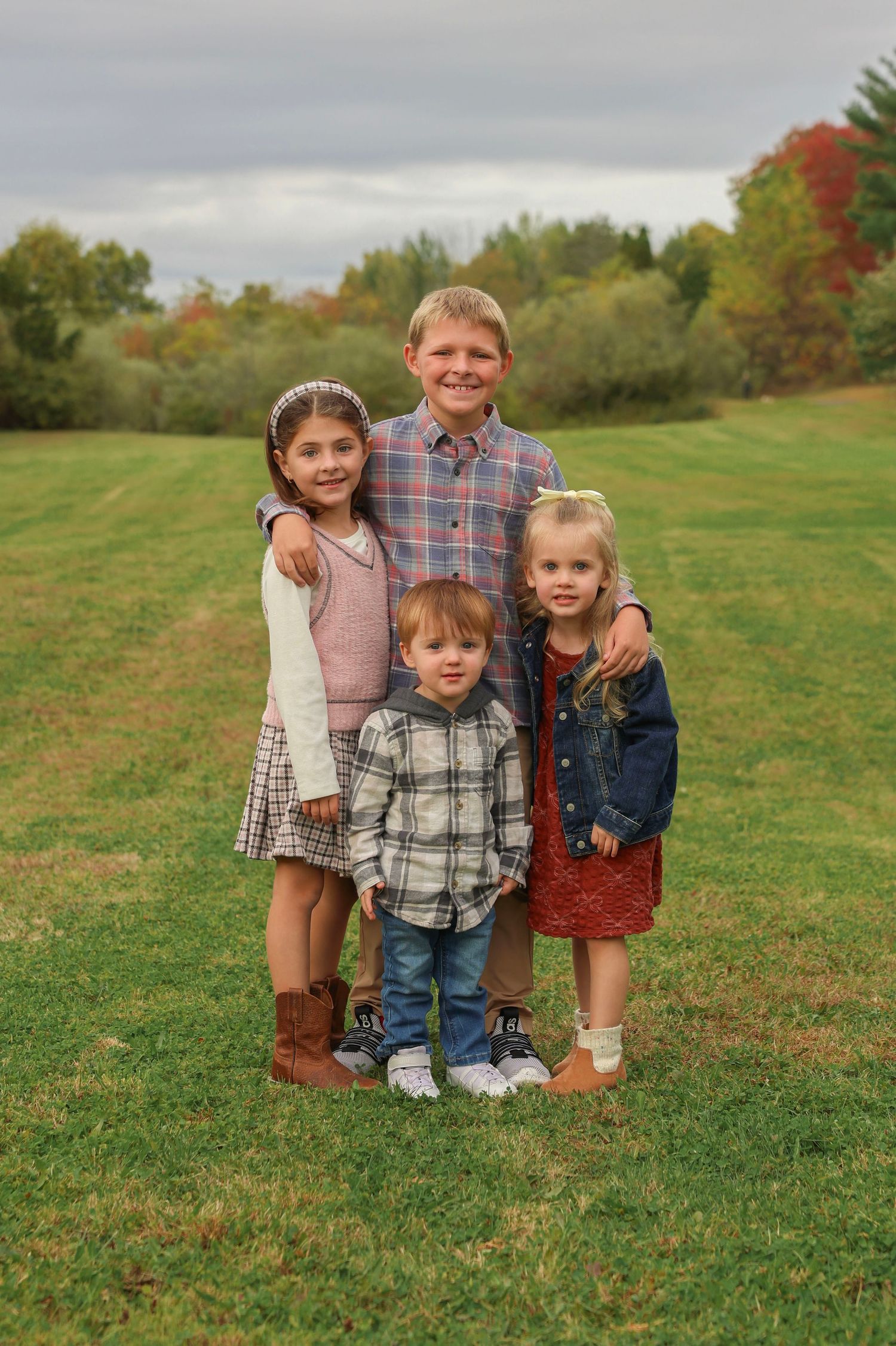 Four children pose together on a green lawn with colorful autumn trees in the background.