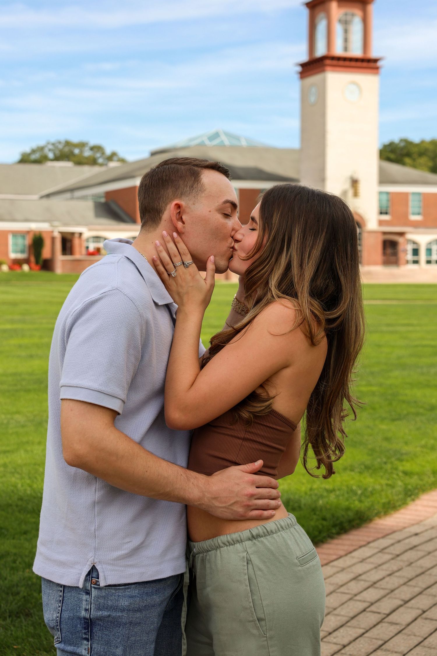 Engaged couple sharing a passionate kiss outdoors on a college campus with a brick clock tower in background.