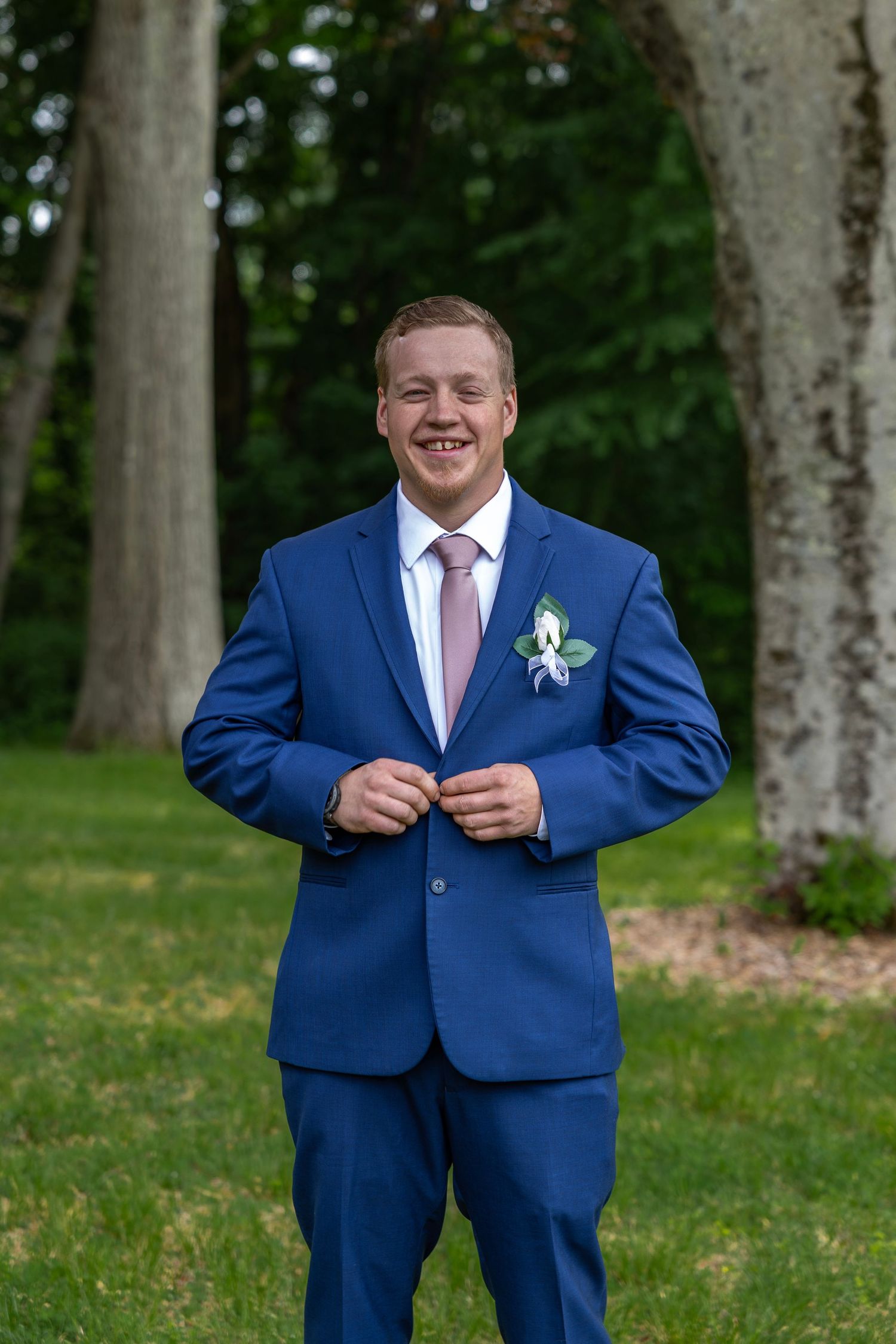Handsome groom in navy blue suit with pink tie and white boutonniere smiling while buttoning jacket outdoors.