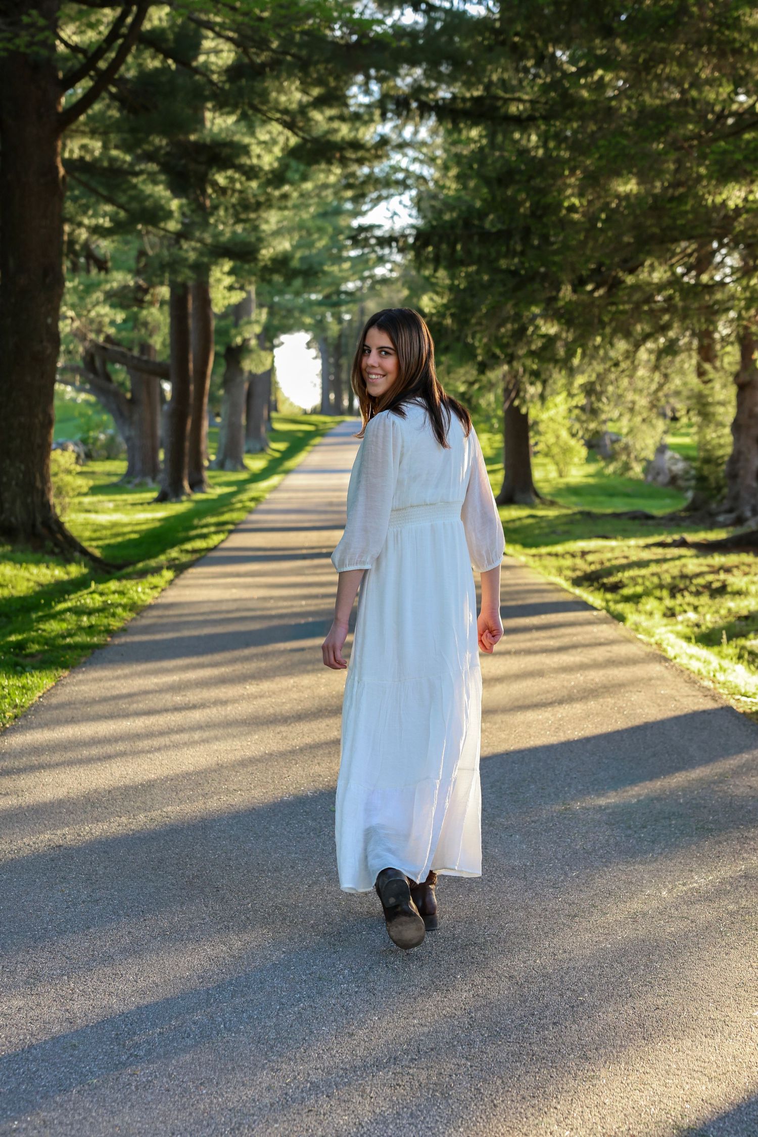 Young woman in white maxi dress walking down a tree-lined road, glancing back with a smile at sunset.