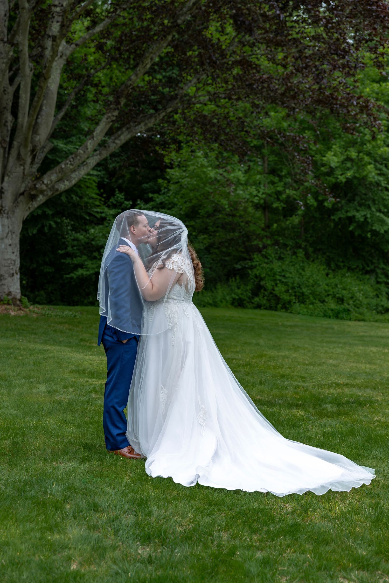 Bride and groom share a kiss hidden under bride's white veil in lush green outdoor garden setting.