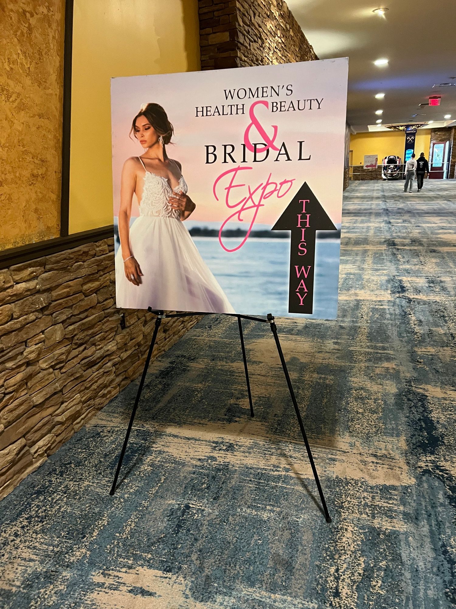 A directional signage board for the Women's Health Beauty and Bridal Expo featuring a bride in a white gown with a this way arrow.