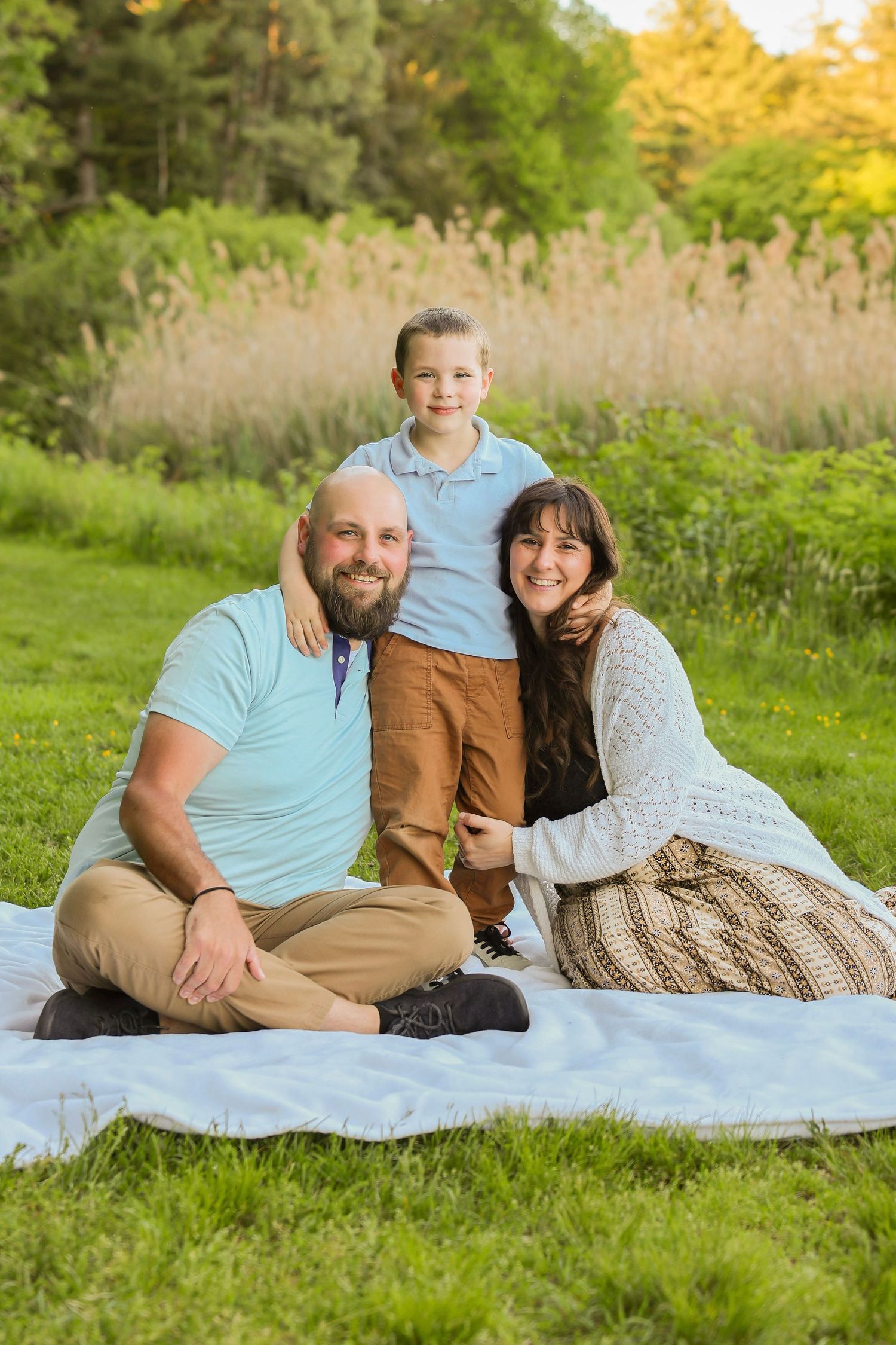 Happy family of three posing on a white blanket in a lush green park during golden hour.