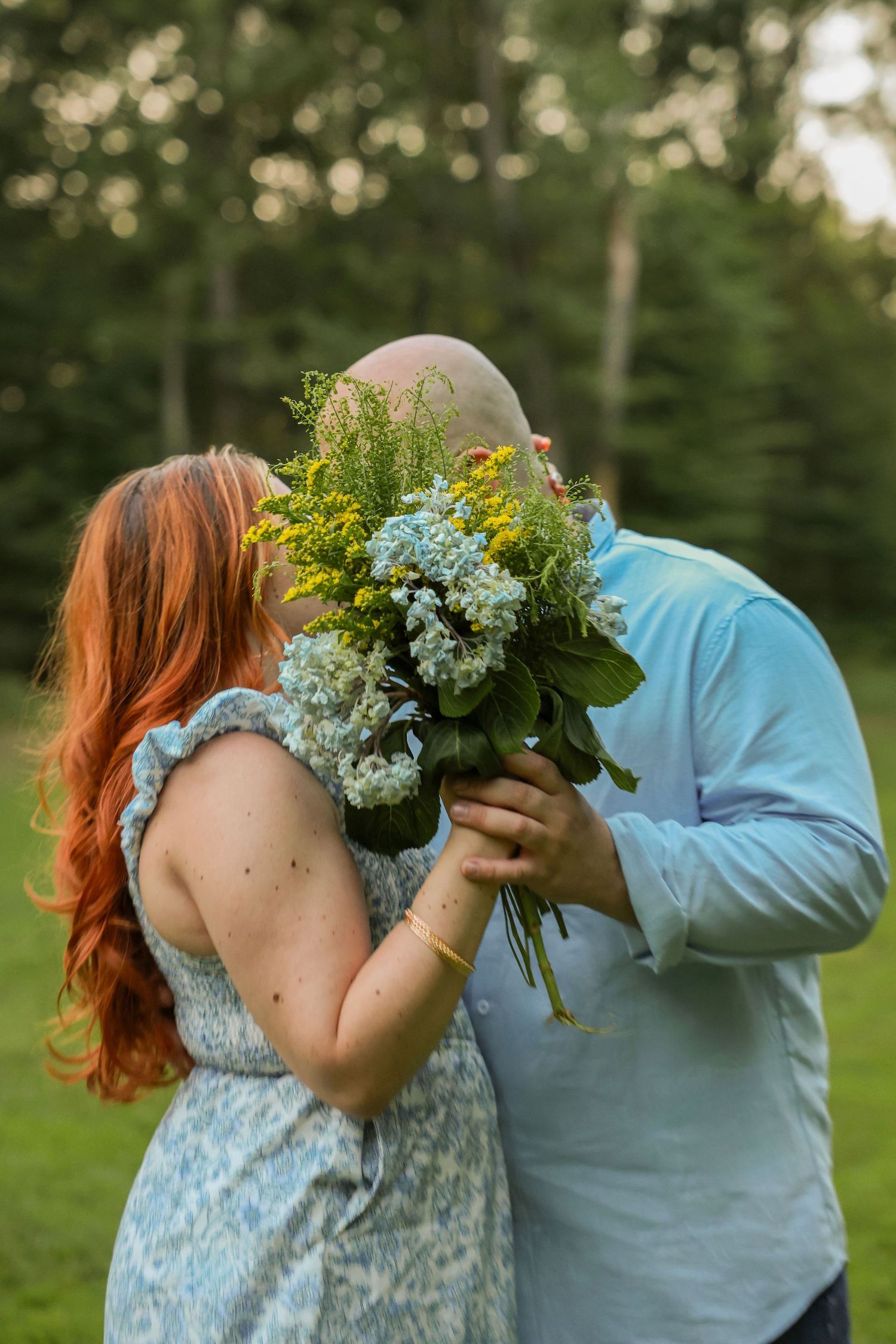 Couple sharing a romantic kiss hidden behind a lush bouquet of blue and yellow wildflowers outdoors.
