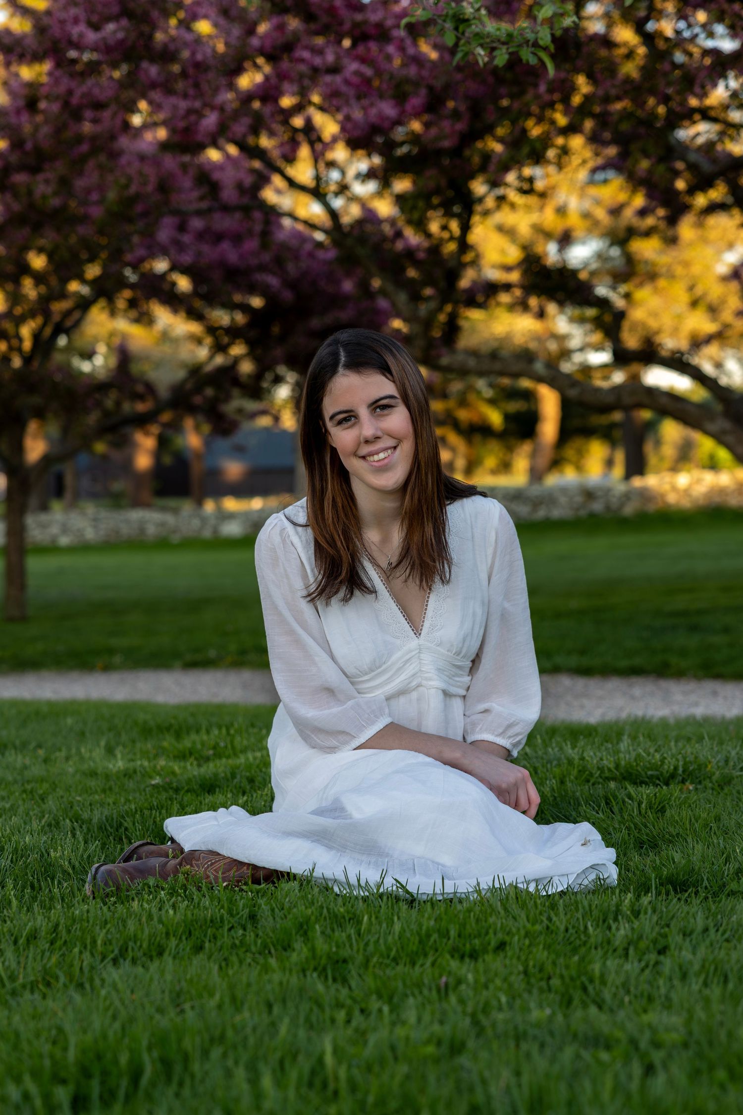 Smiling young woman in white dress sitting cross-legged on grass near pink trees.