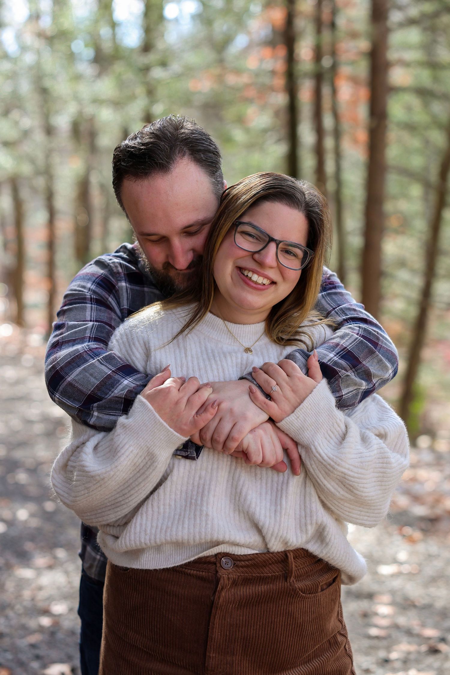 A man hugs a smiling woman from behind on a sunlit forest path during an autumn engagement session.