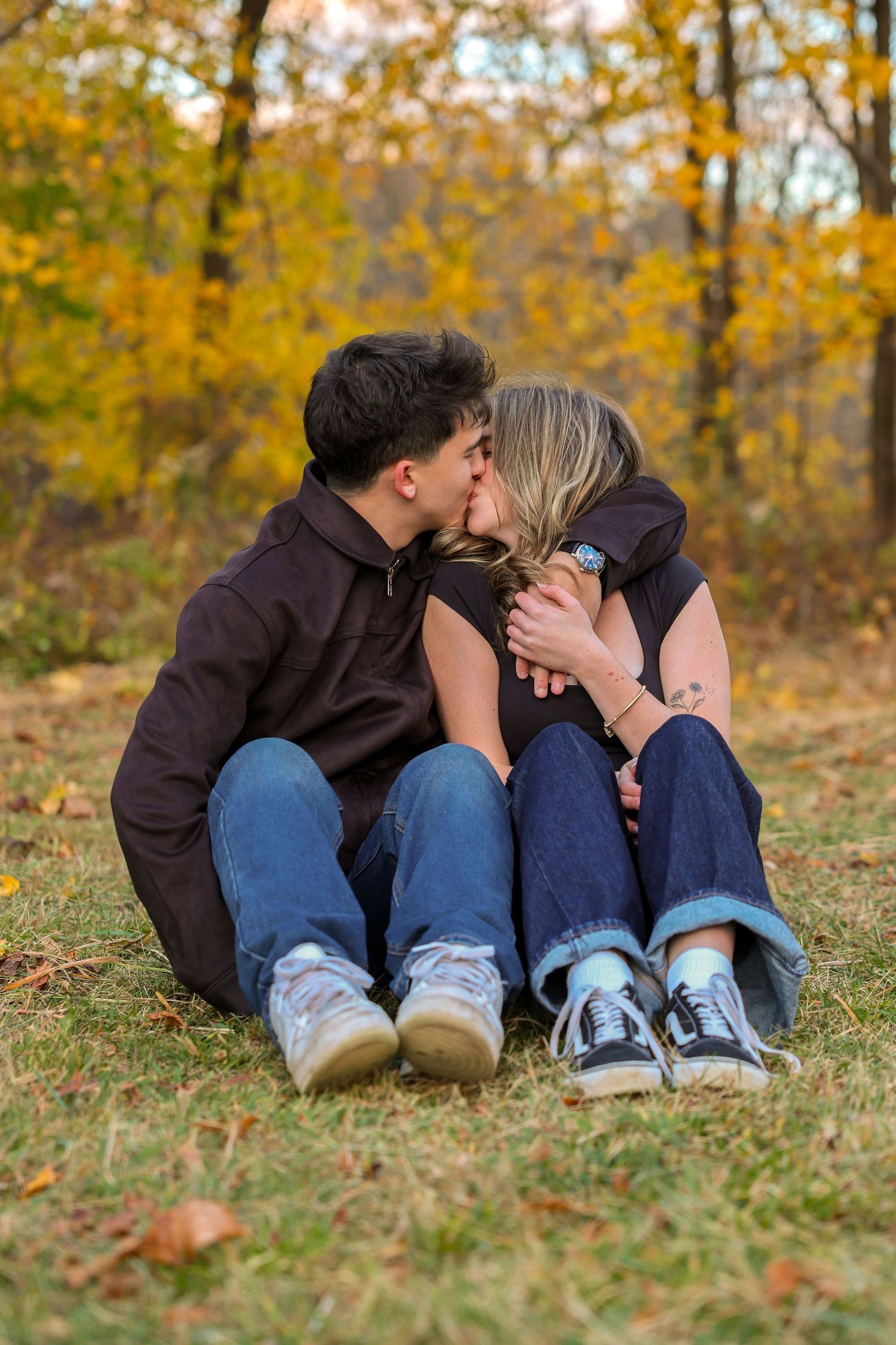 Romantic couple sharing a kiss while sitting on grass surrounded by golden autumn leaves.