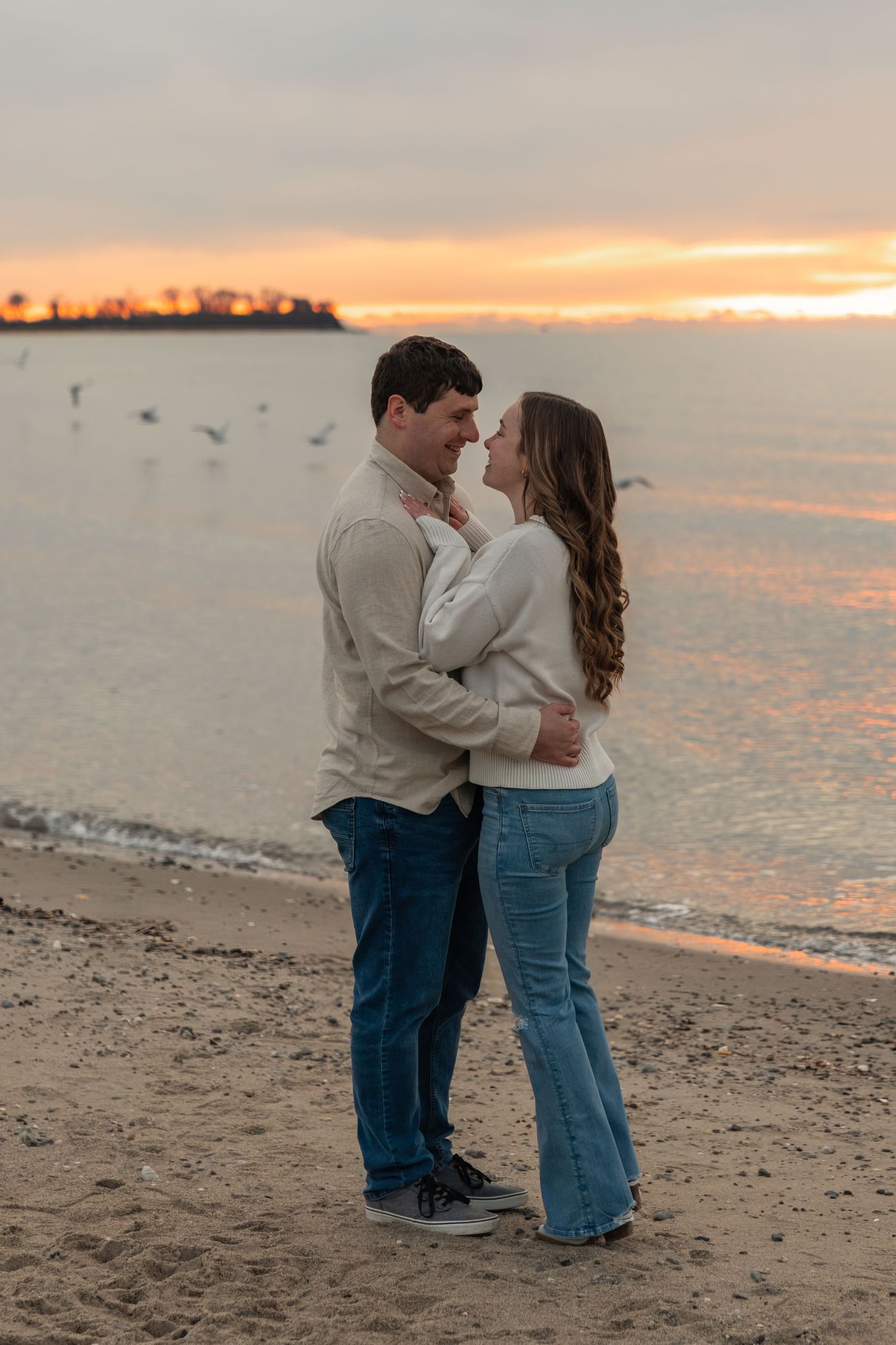 Happy couple embracing on sandy beach with birds flying over colorful sunset sky.