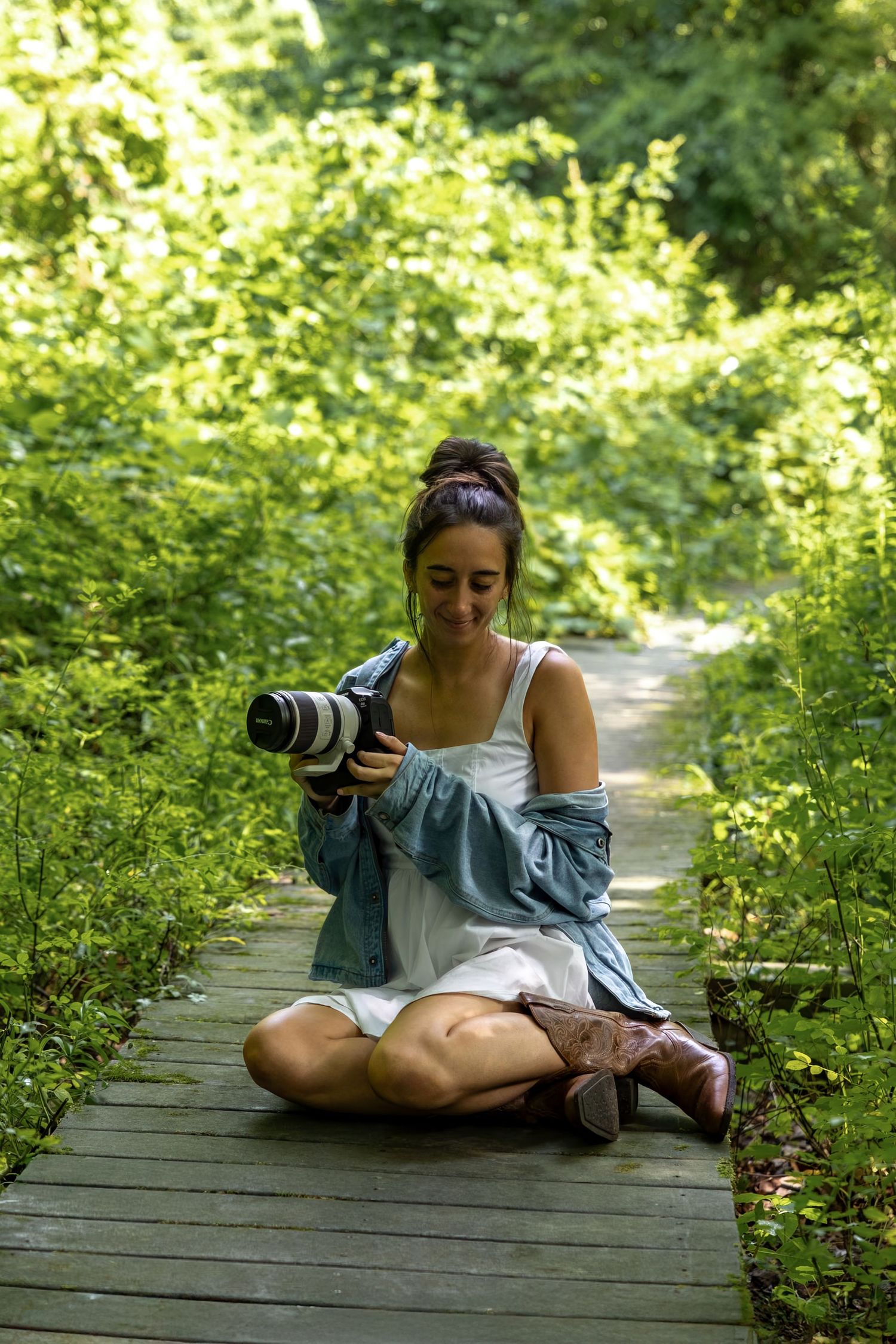 Young woman sitting cross-legged on a wooden path in a forest, reviewing photos on a DSLR camera.