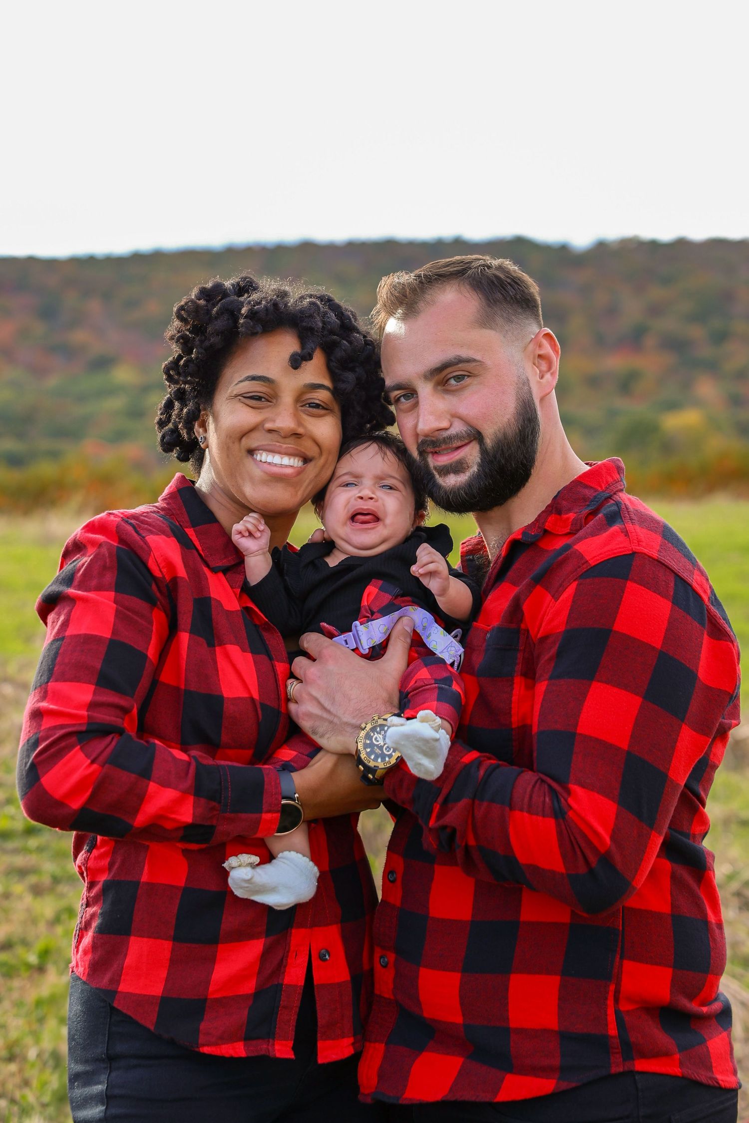 Interracial couple in matching red plaid shirts holding crying newborn baby outdoors with colorful autumn hills behind.