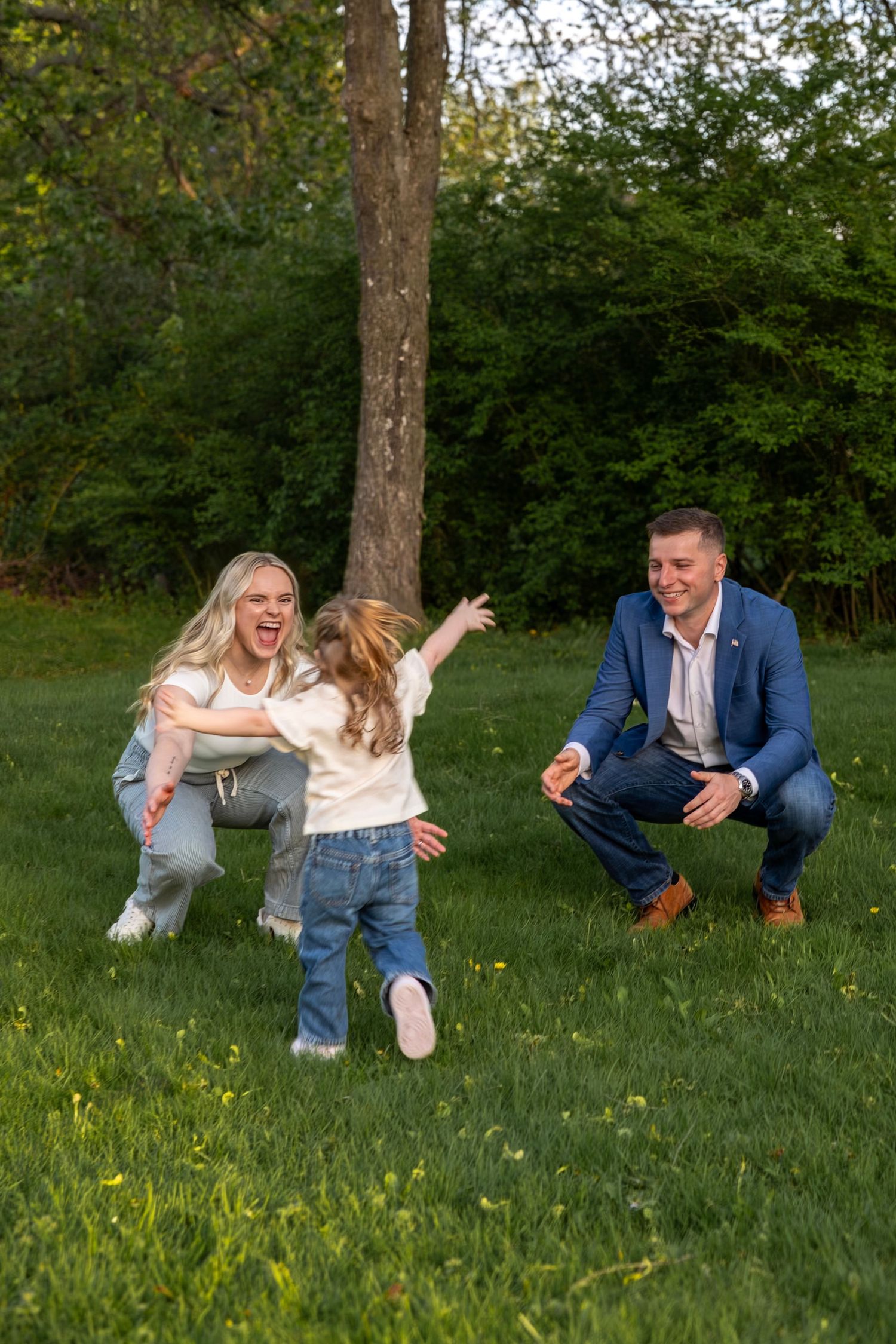 Excited mother and father playing with running toddler on green grass surrounded by trees in park.