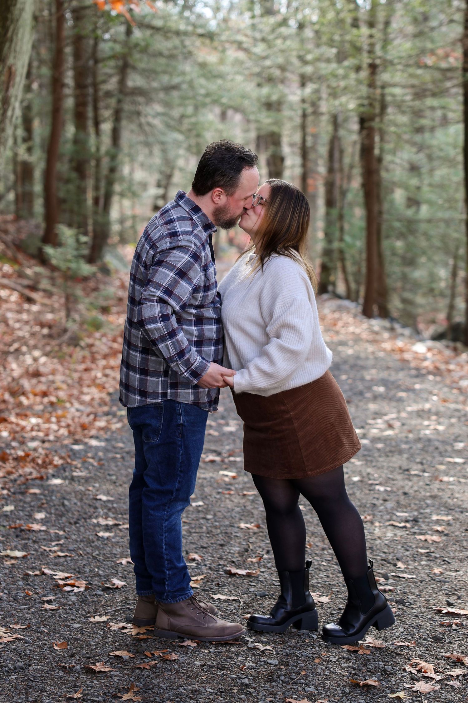 Couple sharing a kiss on a leaf-covered forest path during autumn, dressed in cozy fall outfits.