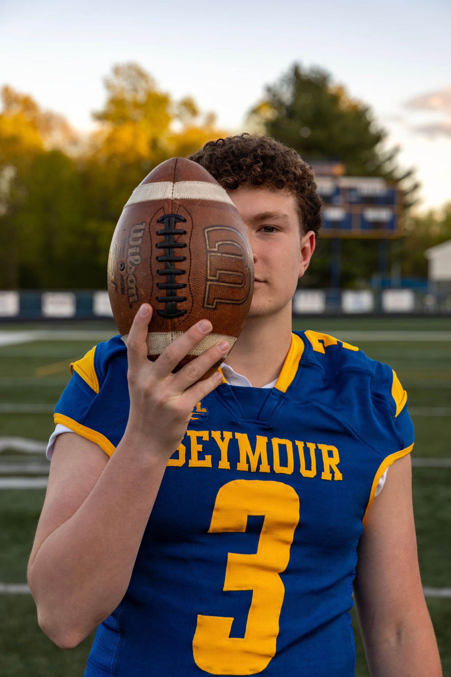 Seymour #3 football player holding Wilson football up to face on field at dusk.