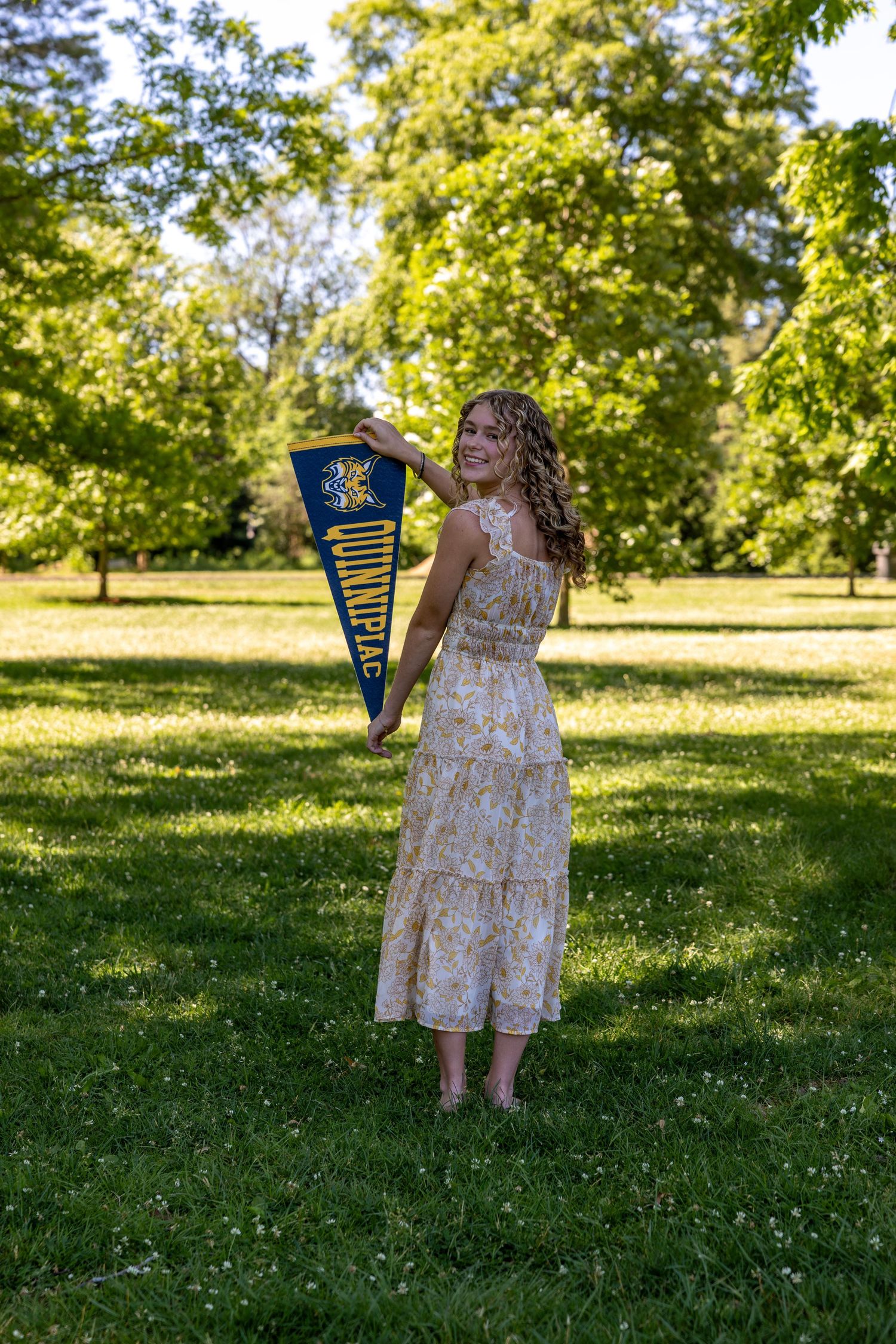 Young woman in floral dress holding a Quinnipiac University pennant flag while posing in a green park.