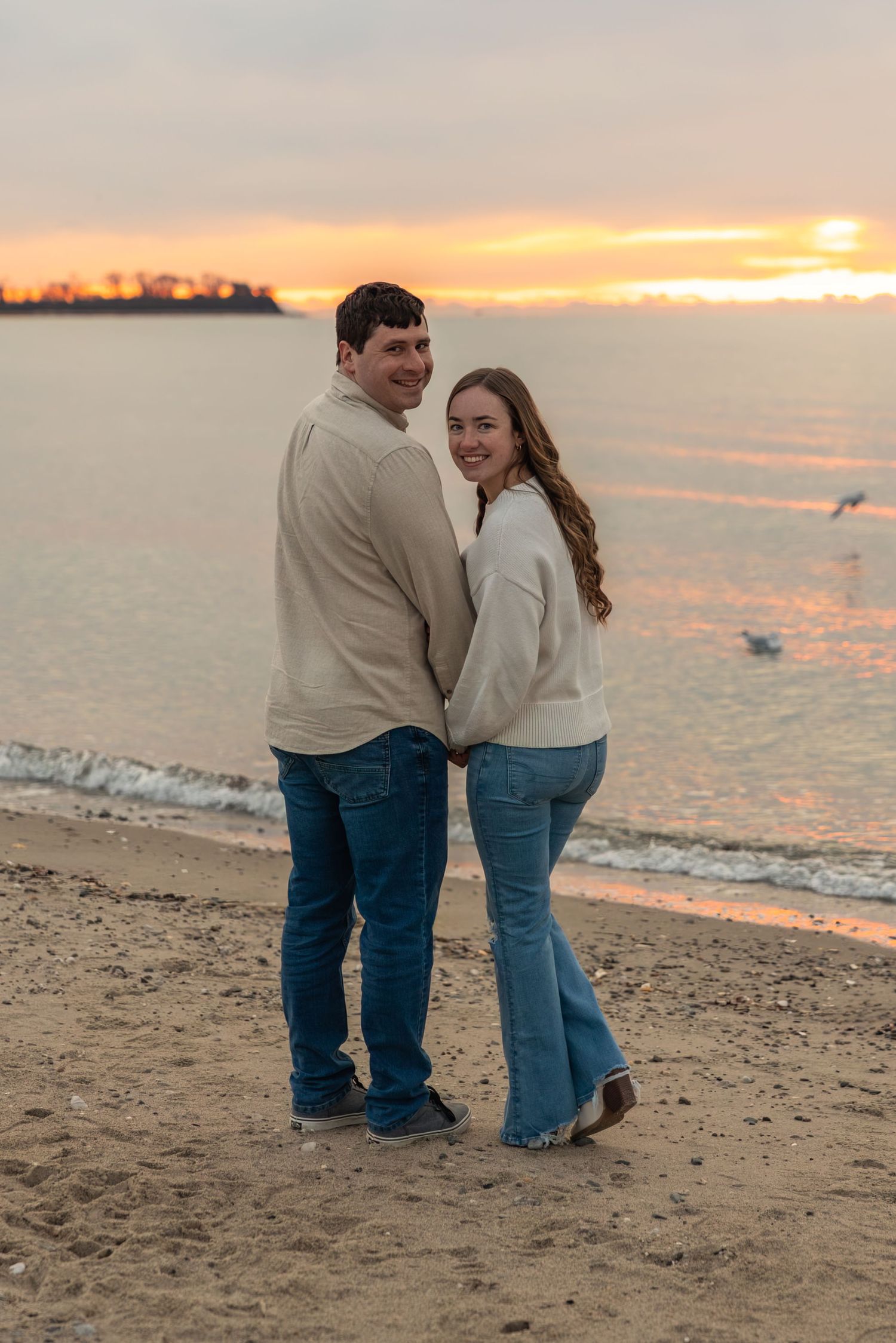 Happy couple holding hands on a beach, smiling at camera during golden sunset.