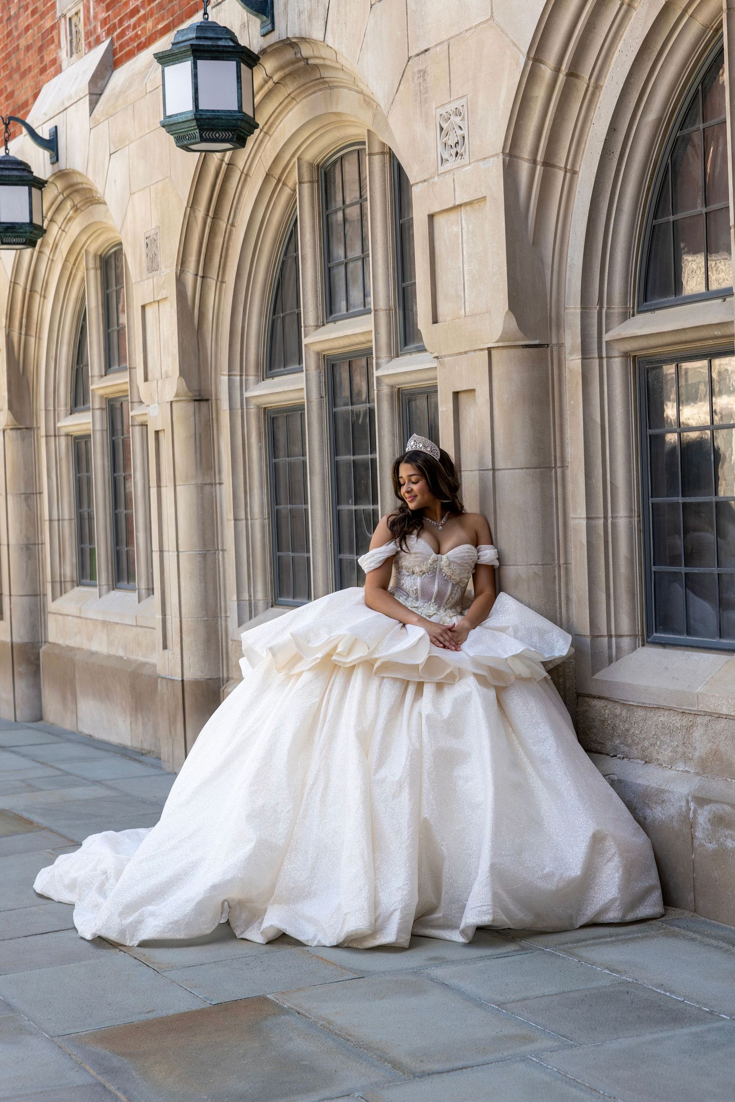 Elegant quinceañera in layered white ball gown leaning against stone archway.