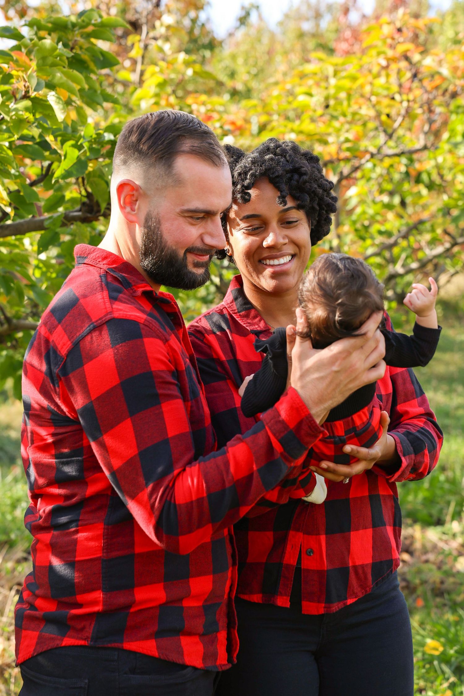 Happy interracial family in matching red plaid shirts cuddling newborn baby outdoors in lush green autumn orchard.