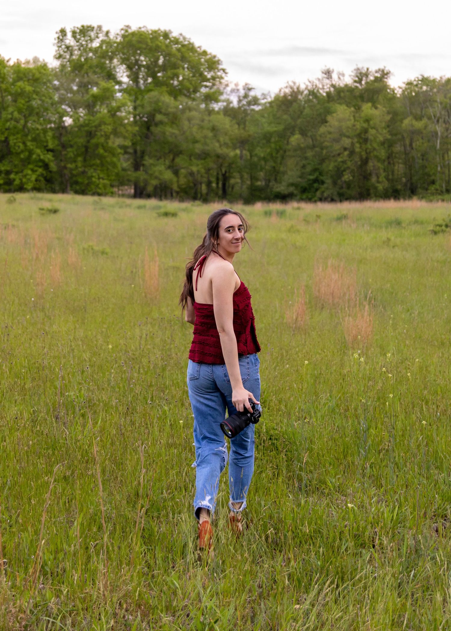 A young woman in a red top and jeans walks through a lush green meadow, glancing back with a smile.