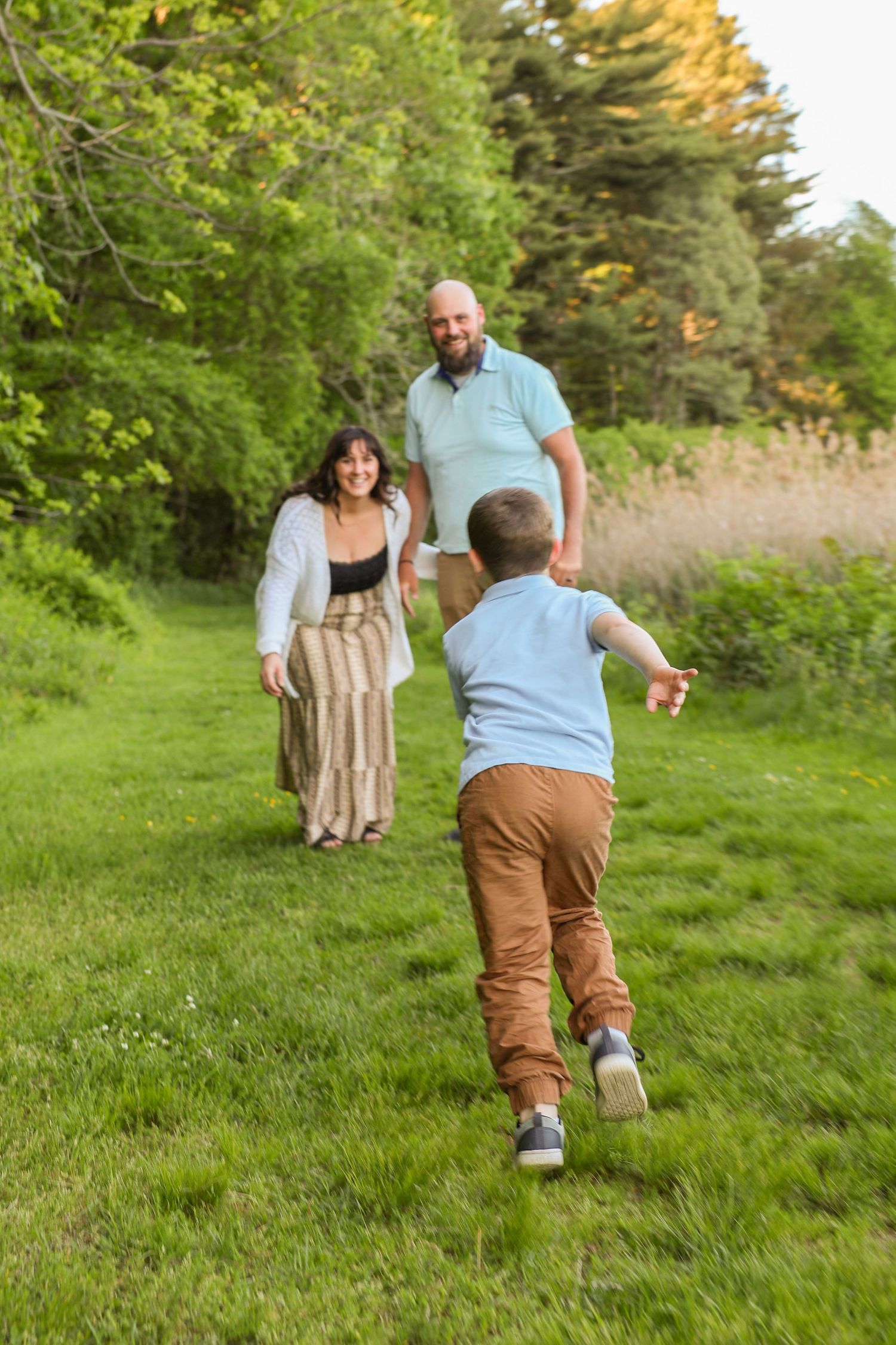 A young boy runs toward his smiling parents across a lush green lawn surrounded by trees.