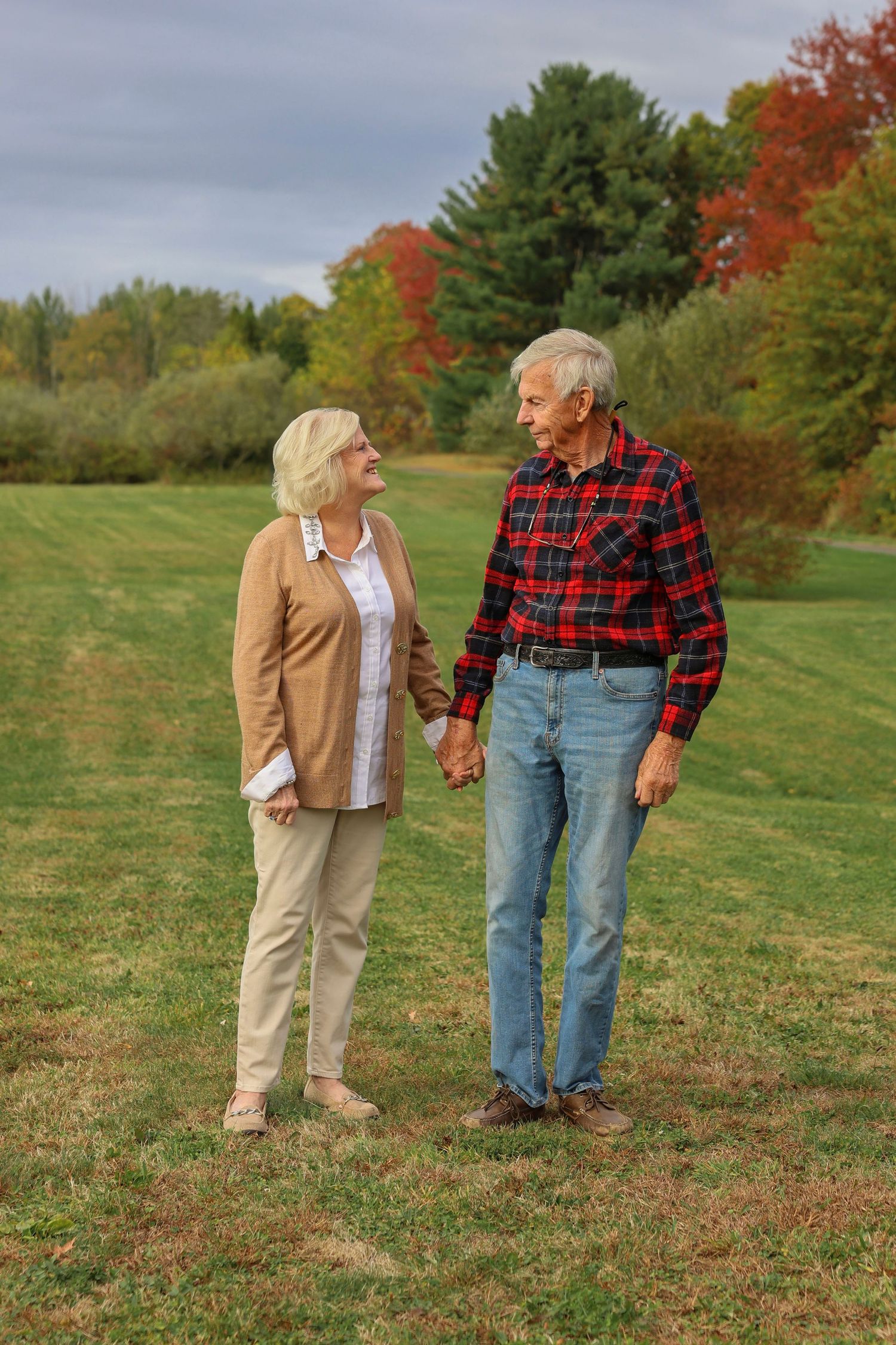 Senior couple holds hands while walking through colorful autumn foliage in outdoor portrait.