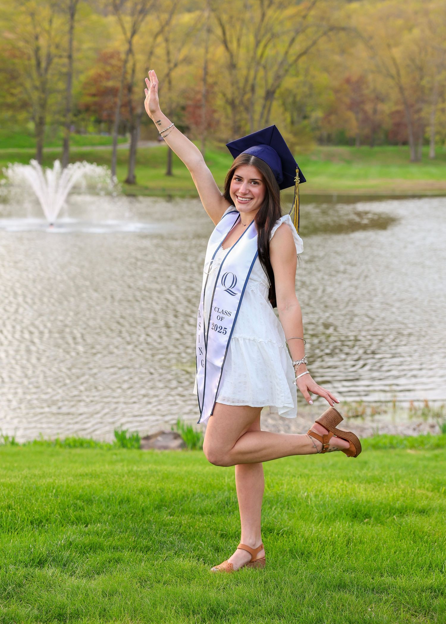 Joyful nursing graduate in cap and white dress posing with raised arm by campus pond.