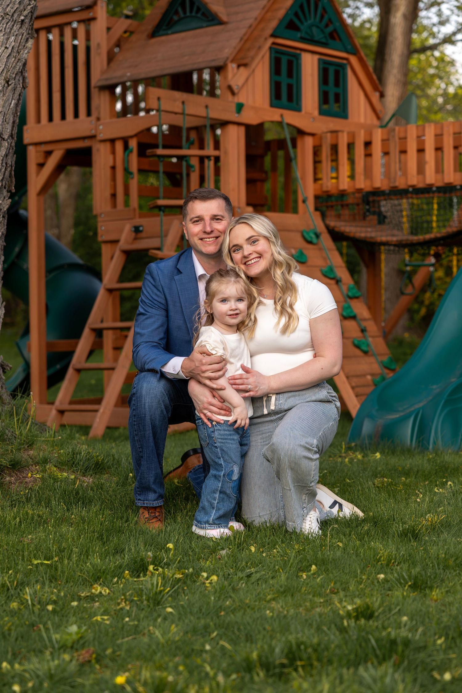 Happy family of three poses together on green grass in front of a large wooden backyard playset.