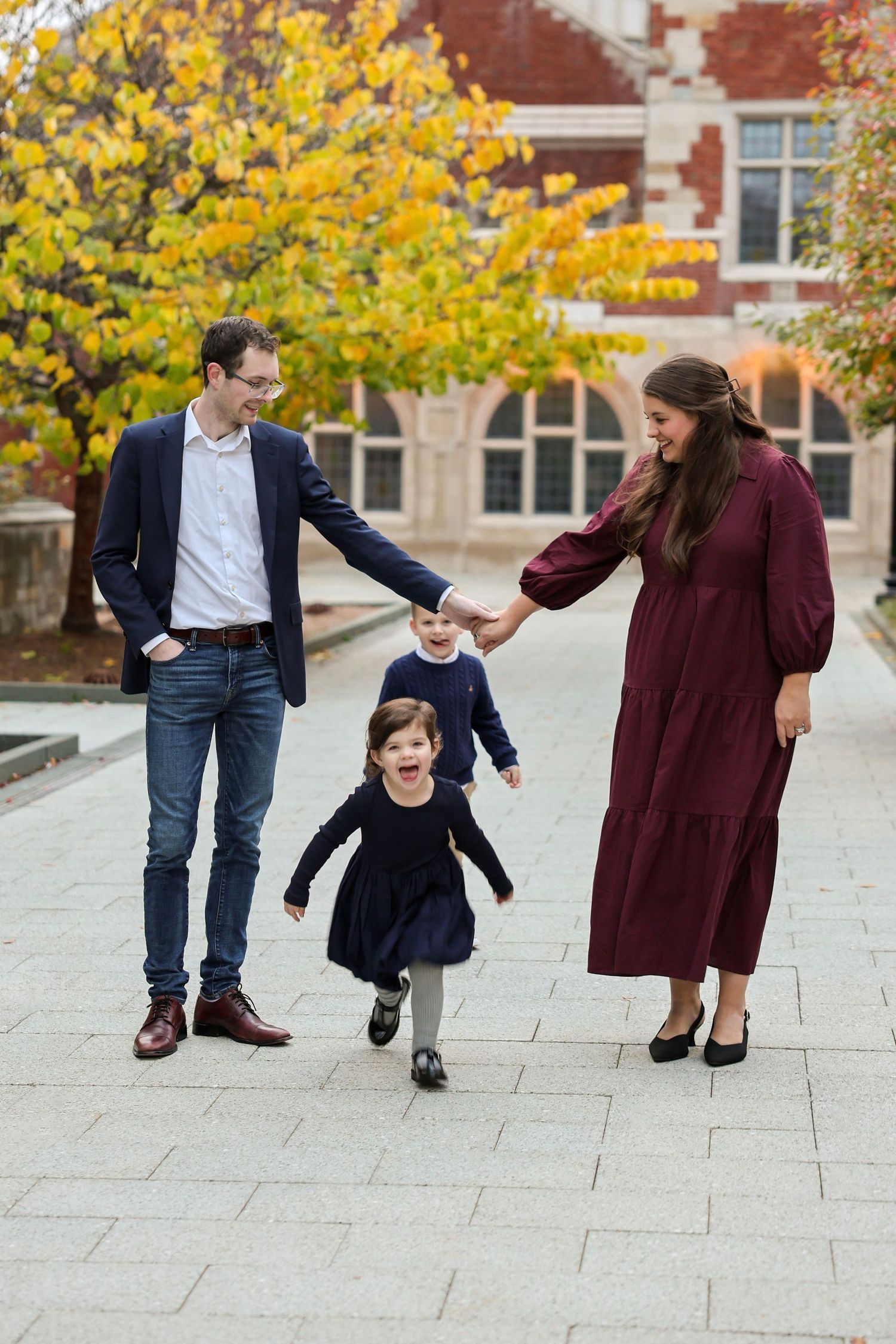 Joyful family portrait with parents and two children playing on a scenic brick pathway amid vibrant fall foliage.