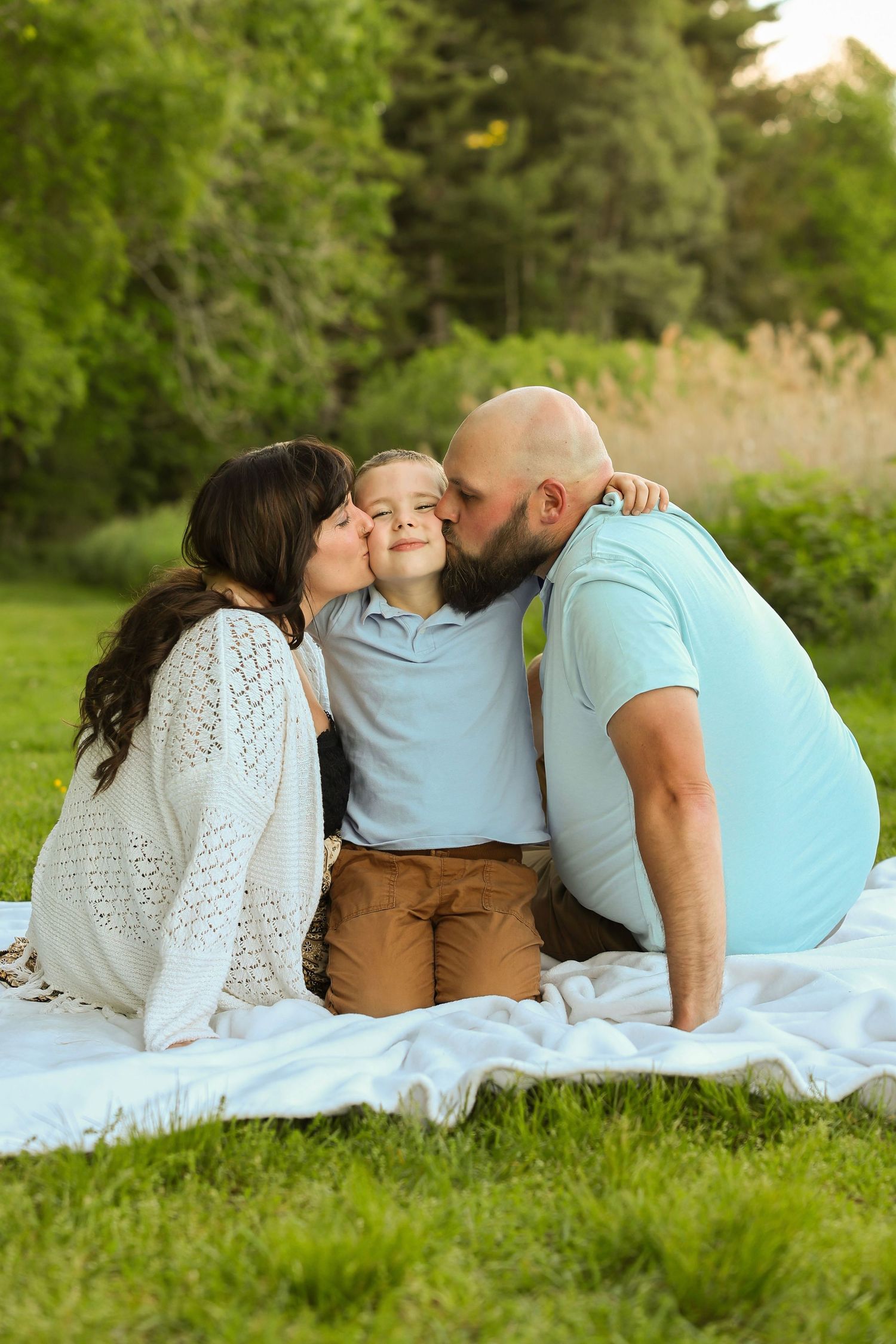 Two parents lovingly kiss their smiling young son on the cheeks while sitting on a blanket outdoors.