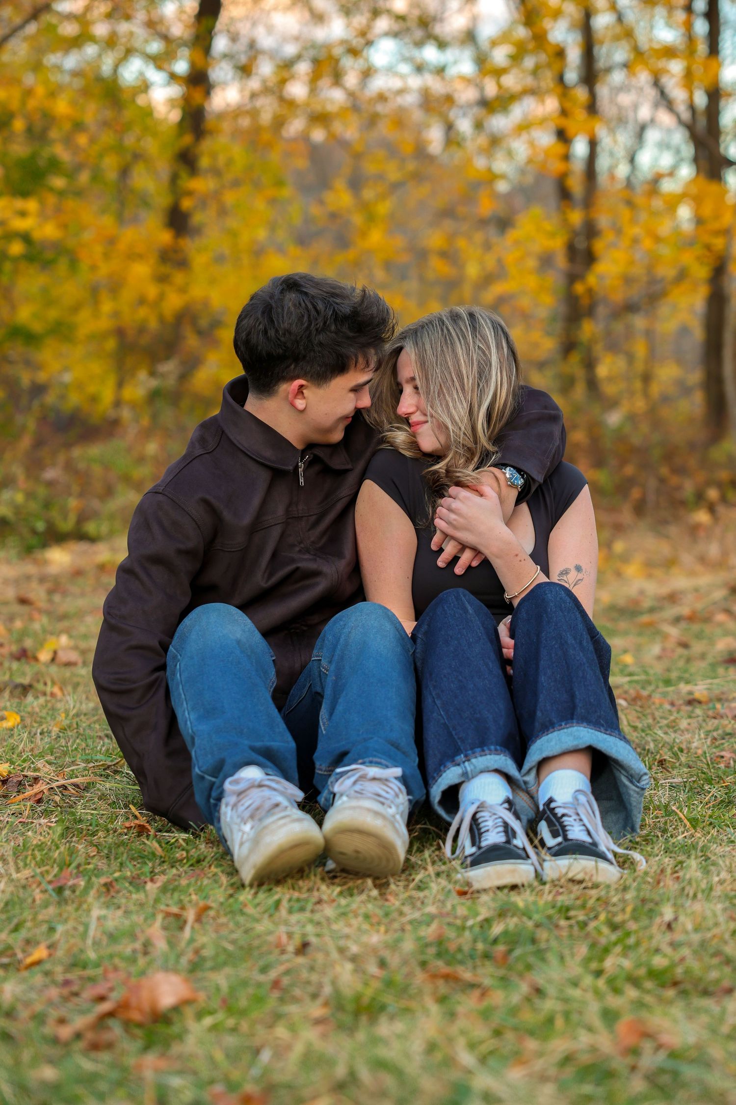 A young couple sitting closely together on grass, sharing a tender moment surrounded by fall foliage.